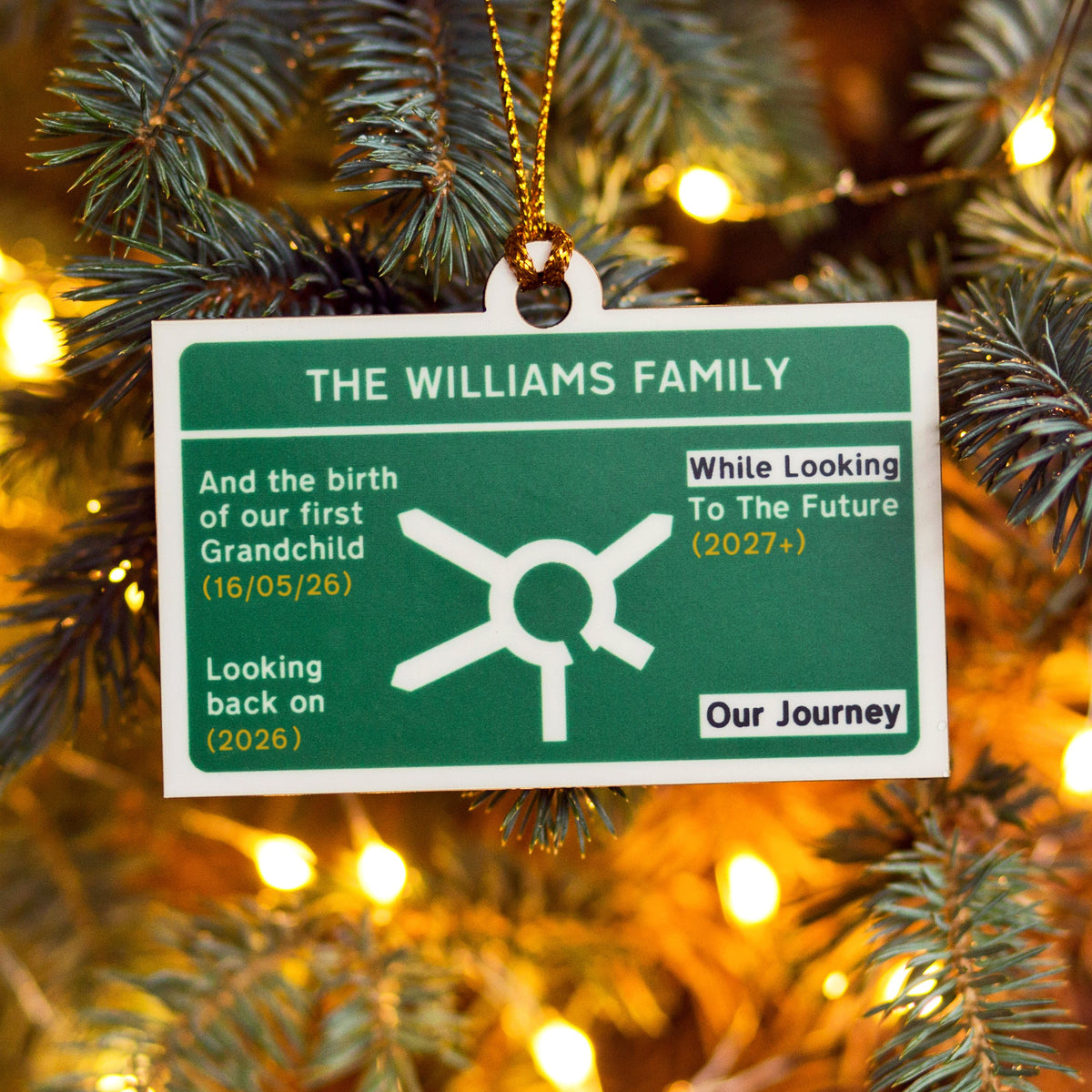 A personalised family Christmas ornament hanging on a Christmas tree. The ornament is a white rectangular plaque with a dark green background designed to look like a motorway road sign and text reading 'THE WILLIAMS FAMILY' and 'Our Journey' alongside a simple white line drawing.