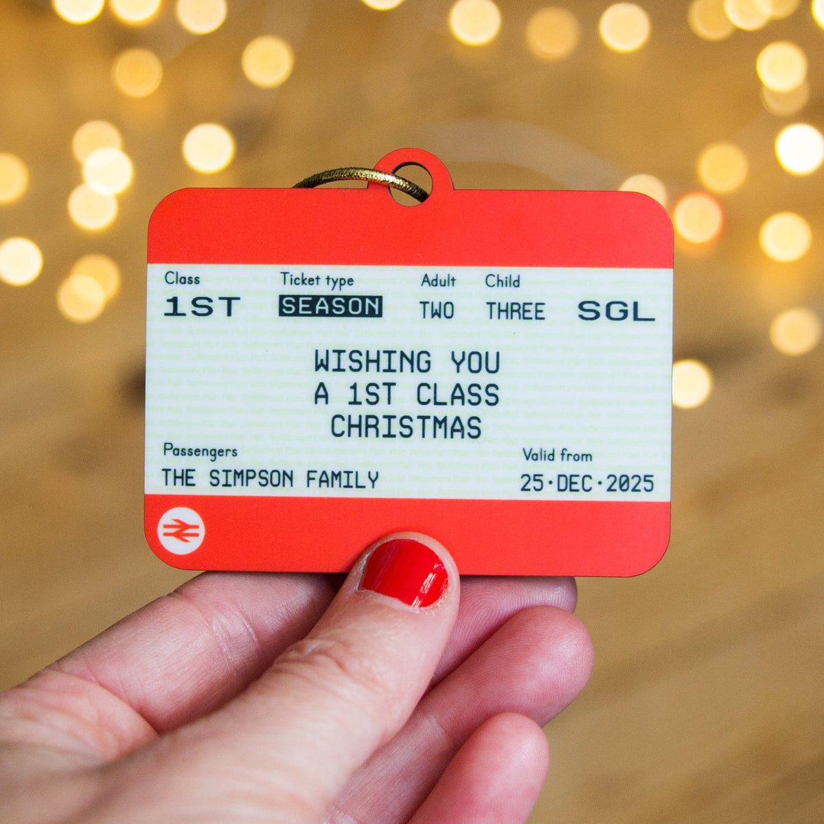 Hand holding a Christmas-themed train ticket ornament with festive lights in the background