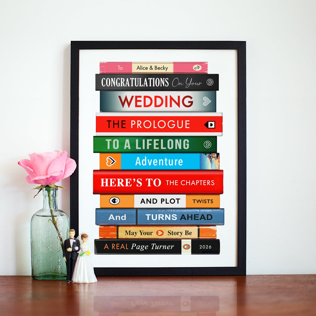 A framed personalised book stack poster with wedding-themed book titles and a 'Congratulations On Your Wedding' message, displayed on a wooden surface next to a pink rose in a glass vase and small wedding figurines against a white wall.