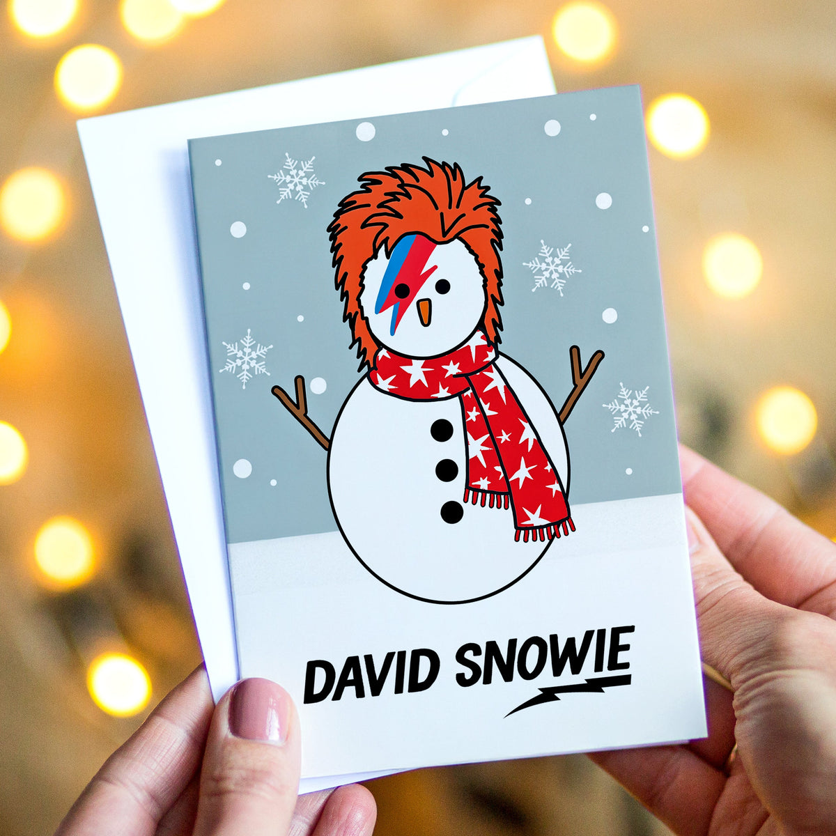 A hand-held greeting card featuring a cartoon snowman styled like David Bowie with a red, white, and blue eye patch, red star scarf, and orange hair, with the text 'DAVID SNOWIE' at the bottom. The card is held by two hands against a warm, blurred background with glowing fairy lights.
