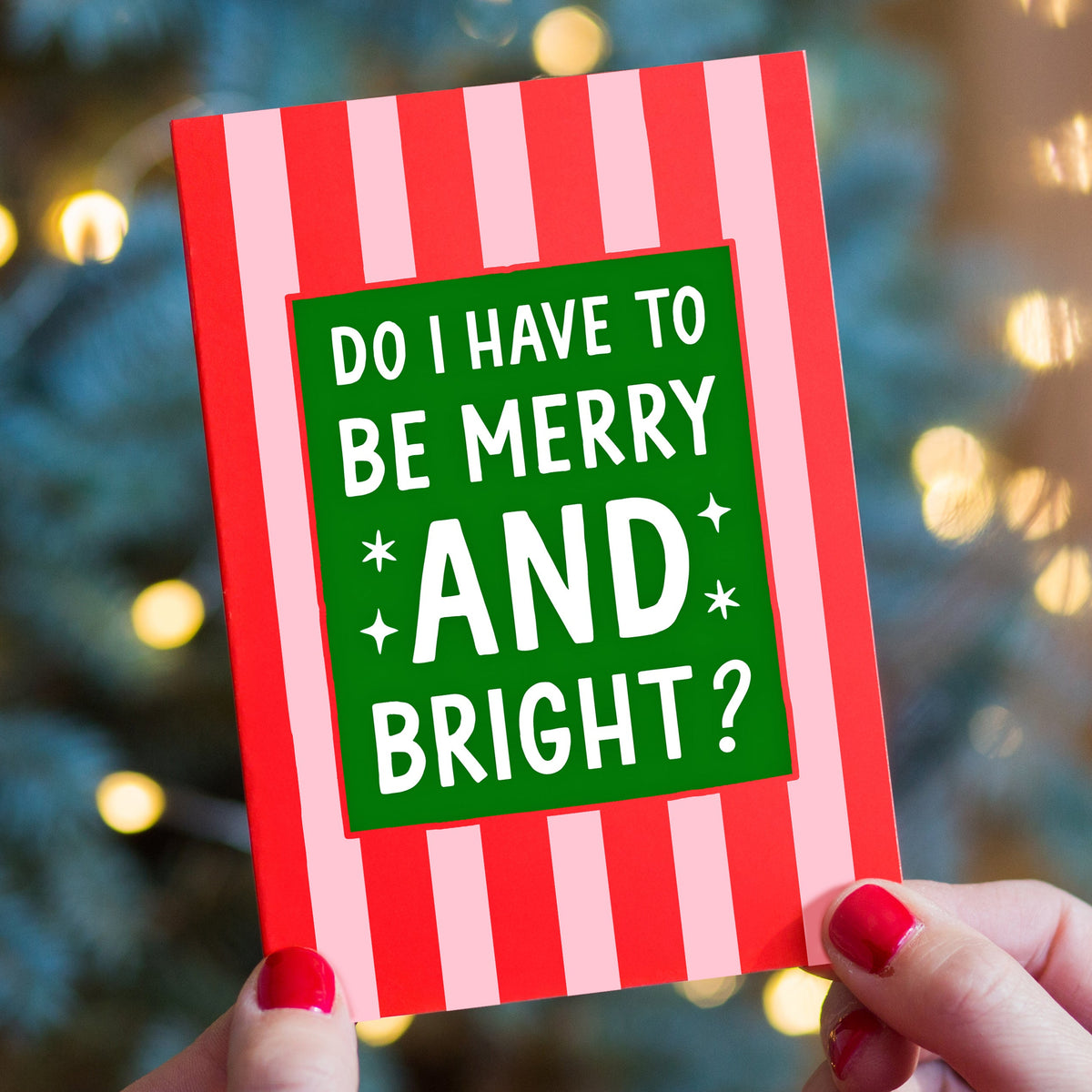 A person holds a red and pink striped Christmas card with the text 'DO I HAVE TO BE MERRY AND BRIGHT?' in white on a green background. The background shows a blurred Christmas tree with warm lights.