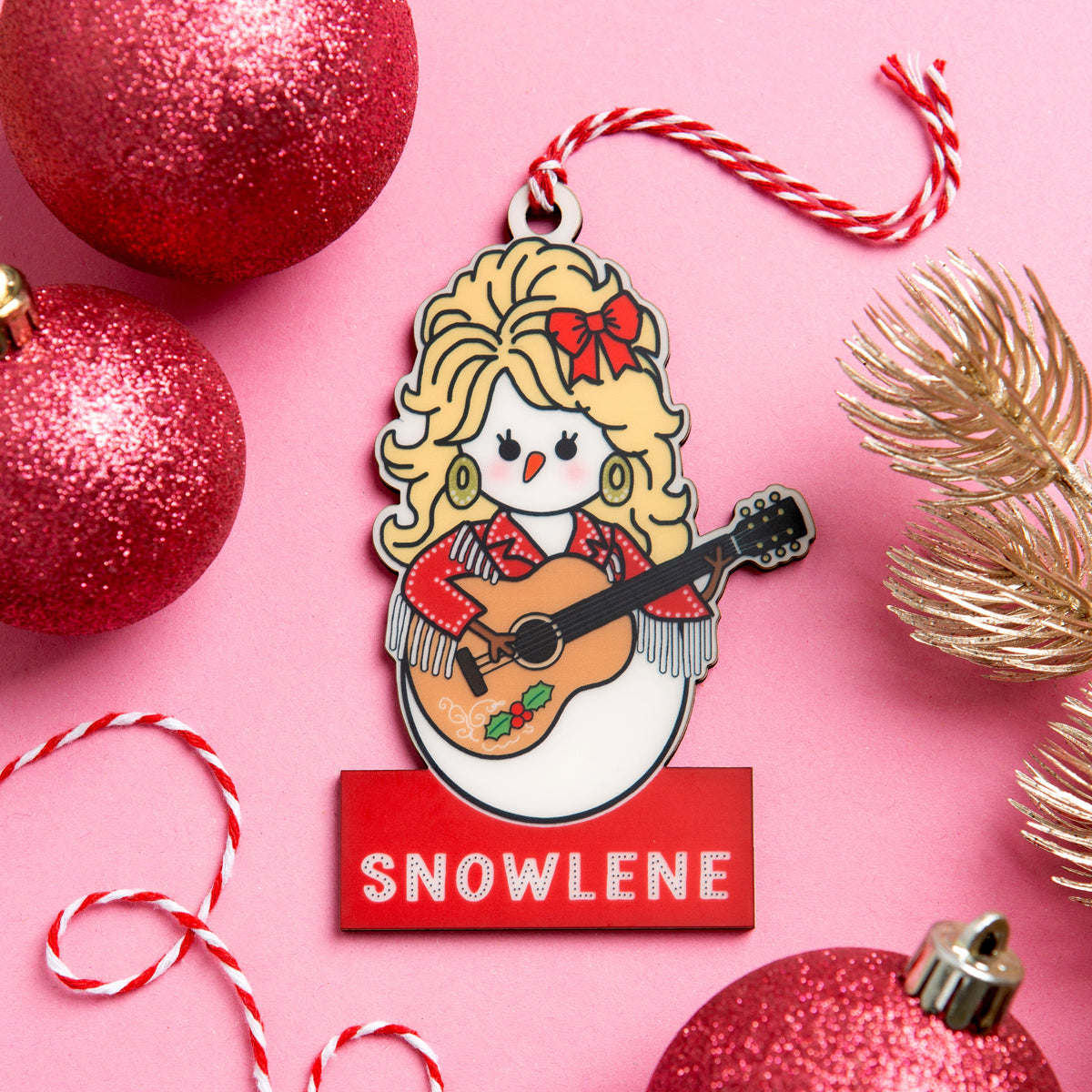 A wooden Christmas ornament featuring a cartoon snowman dressed as dolly parton playing a guitar with the name 'SNOWLENE' on a red bar below. The ornament is styled on a pink background with red glitter ornaments, a gold pine branch, and red-and-white candy canes.