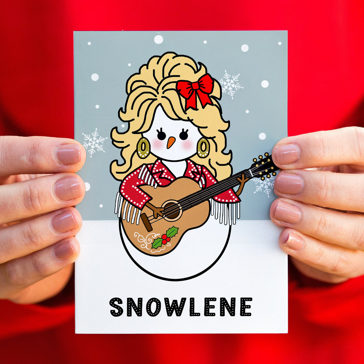 A person holds a Christmas card featuring a cartoon snowman dressed as Dolly Parton playing a guitar, with the word "SNOWLENE" printed below. The background is a bright red fabric, and only the person's hands and torso are visible.