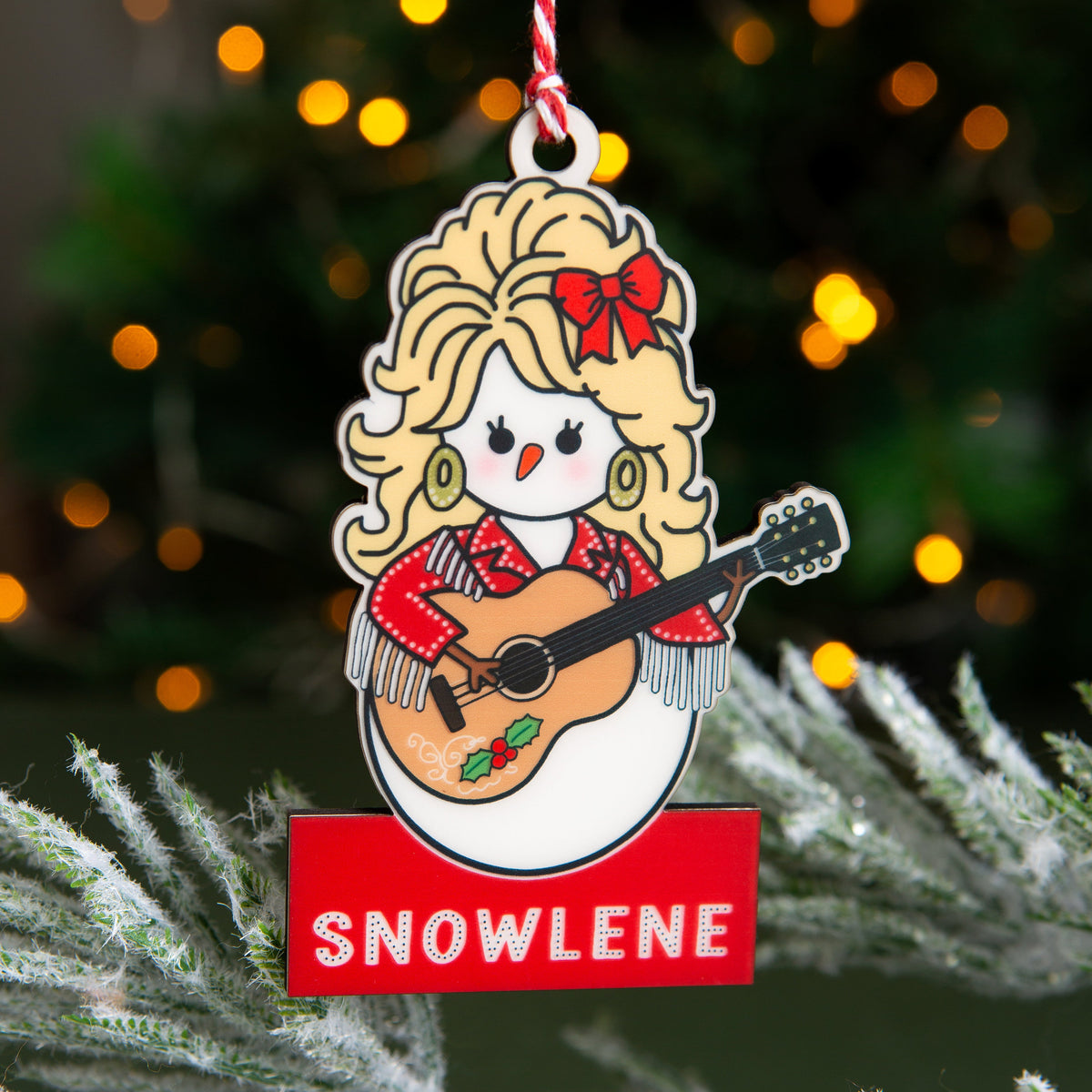 A Christmas ornament shaped like a snowman dressed like Dolly Parton playing an acoustic guitar, hanging from a red and white striped string. The ornament features the name 'SNOWLENE' in white text on a red banner at the bottom.