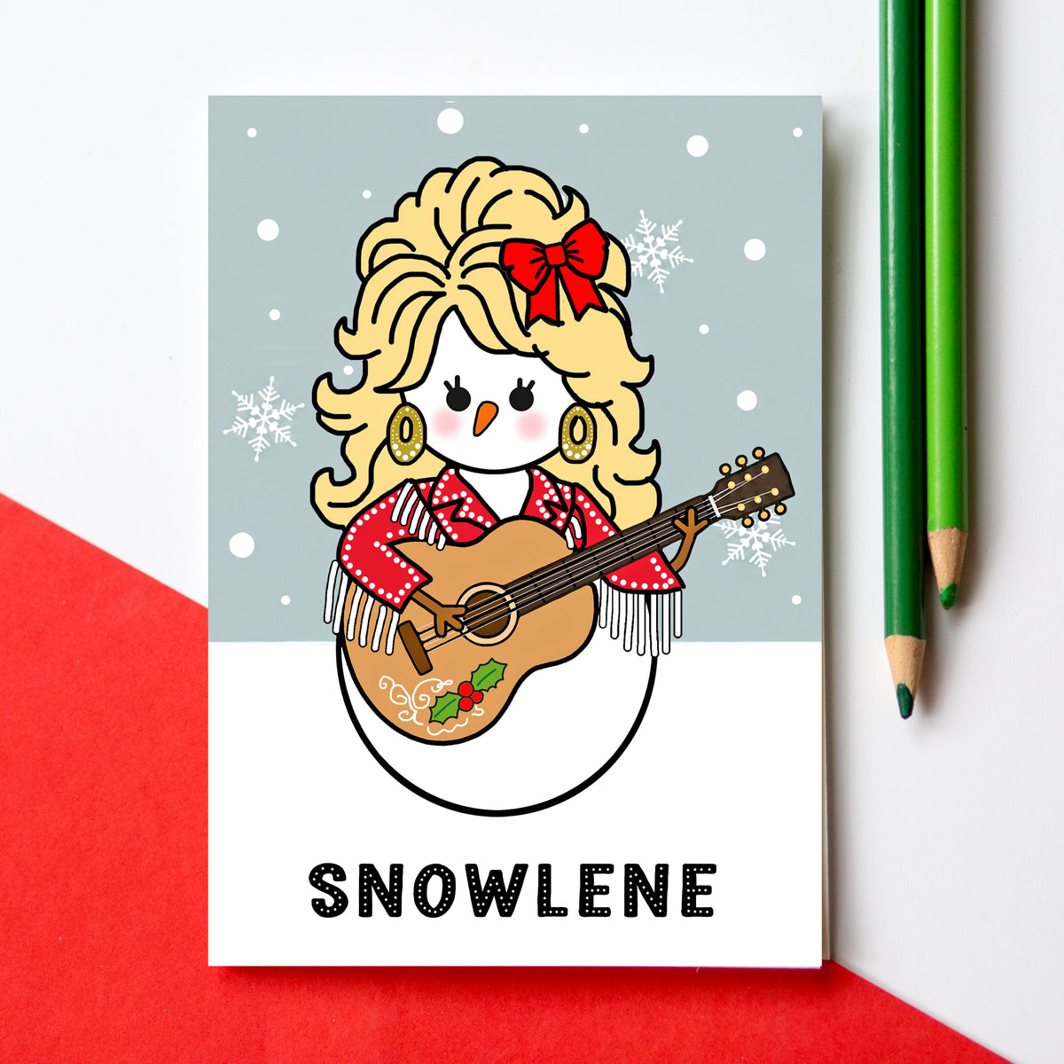 A Christmas  card featuring a cartoon snowman dressed like Dolly Parton with blonde hair, a red bow, and a guitar, with the name 'SNOWLENE' printed below. The card is styled on a white surface with a red paper corner and two green pencils.