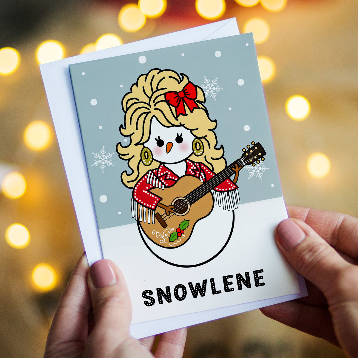 A person holds a custom illustrated Christmas card featuring a cartoon Dolly Parton snowman playing a guitar and the name 'SNOWLENE' at the bottom. The background shows warm, out-of-focus string lights.