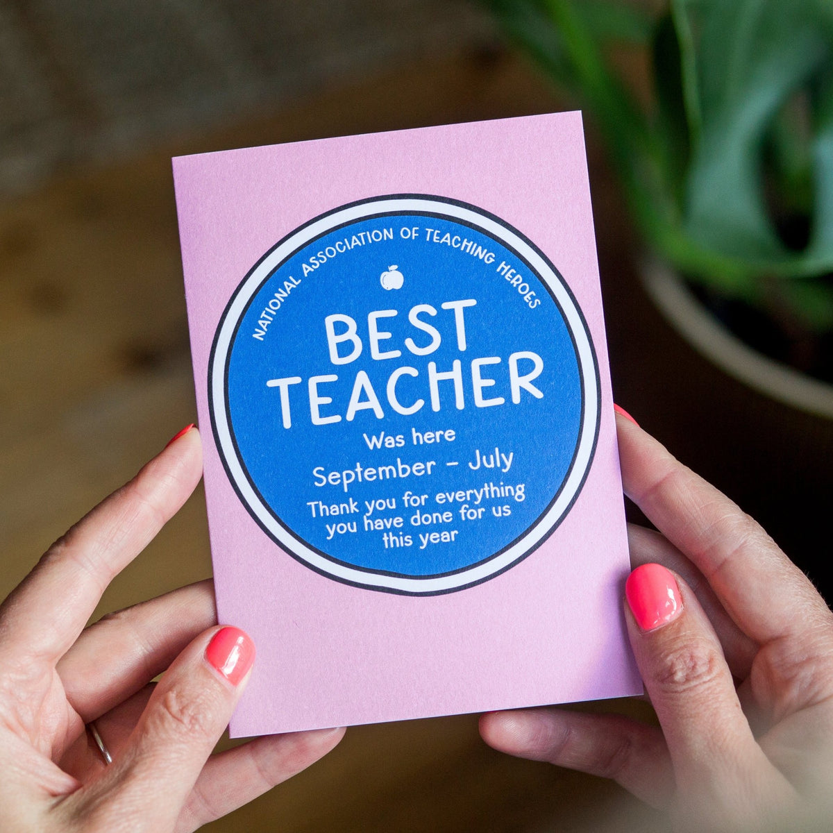 Card with 'Best Teacher' text held by hands with pink nail polish, blurred plant in background