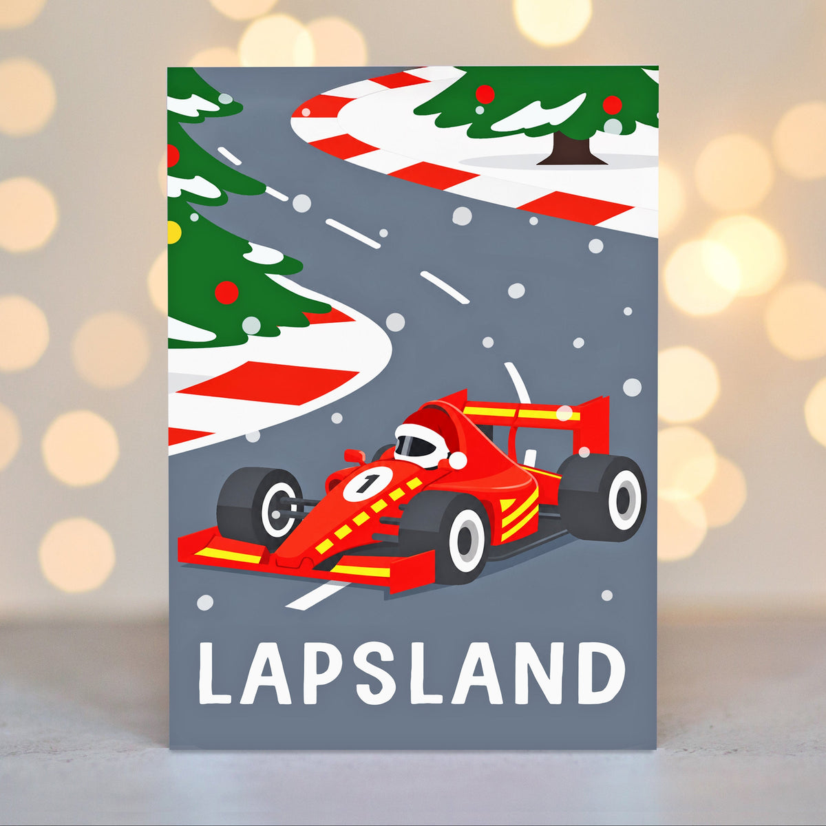 A rectangular greeting card featuring a festive Christmas-themed racing scene with a red race car on a gray road, holiday trees, candy cane stripes, and the word 'LAPS LAND' at the bottom, photographed against a softly blurred warm bokeh background.