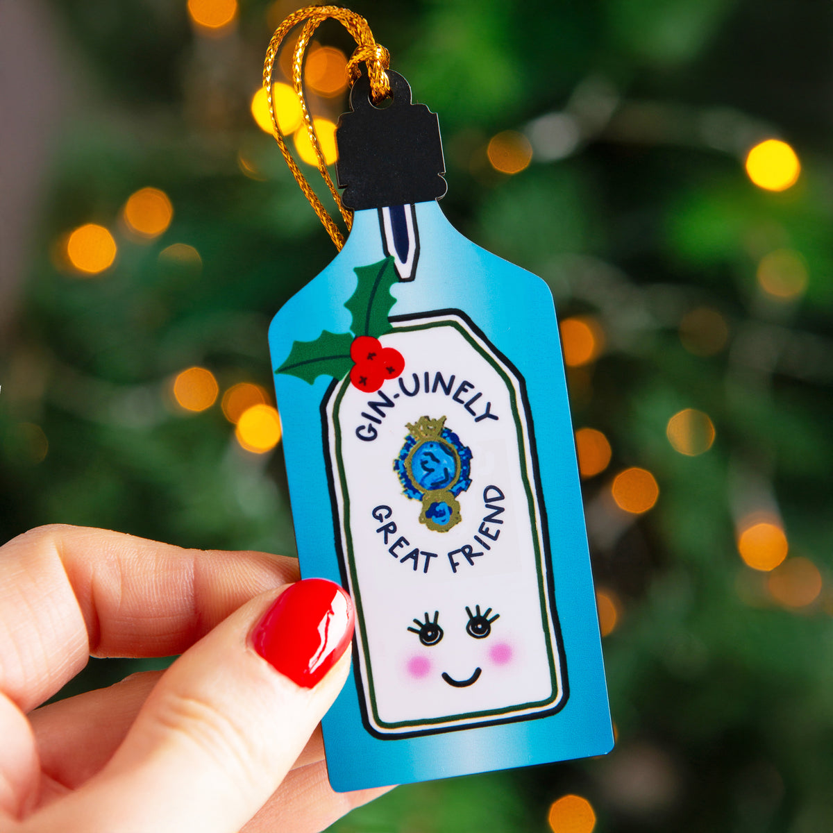 A hand with red nail polish holds a blue Christmas ornament shaped like a gin bottle. The ornament features a white label with the text 'GIN-UNELY GREAT FRIEND', a blue medallion, holly leaves, red berries, and a cute smiling face.