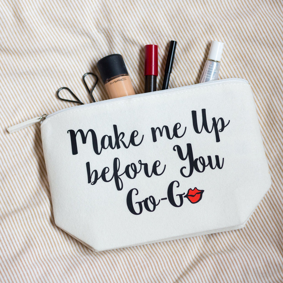 A white cosmetic pouch with black text reading 'Make me Up before You Go-Go' and a small red lips graphic, shown on a beige striped fabric background with makeup items sticking out of the top.