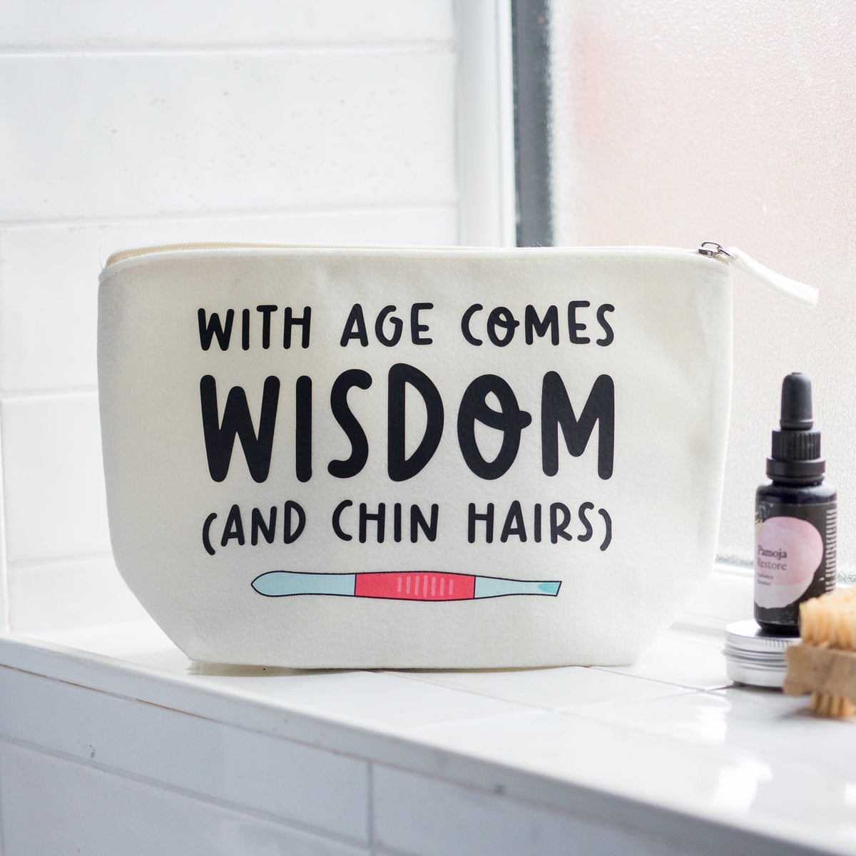 An off-white canvas cosmetic pouch with black text reading 'WITH AGE COMES WISDOM (AND CHIN HAIRS)' and a small illustrated eyebrow product, shown on a white tiled windowsill next to a small bottle and a wooden brush.