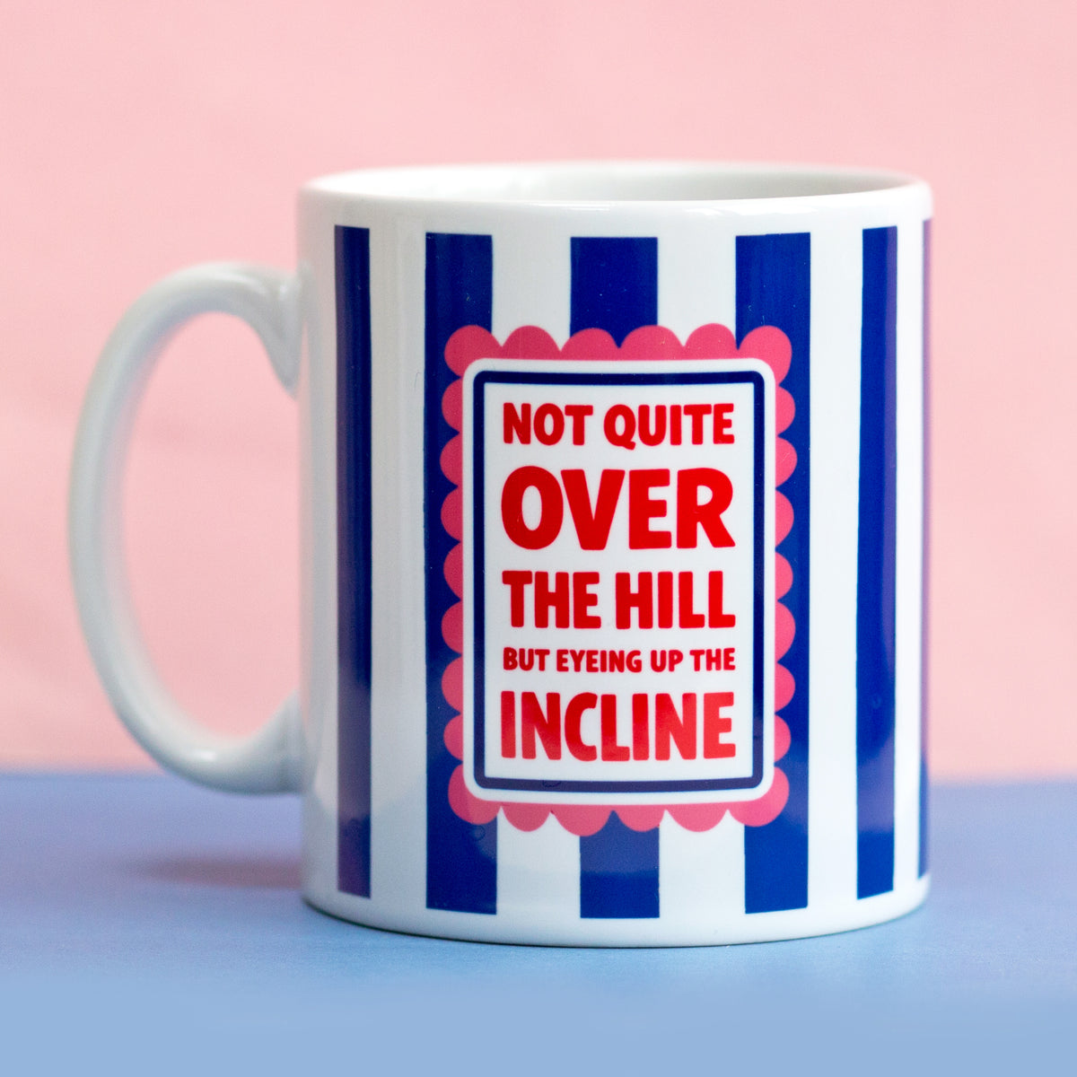 mug with blue and white stripes that says not quite over the hill but eyeing up the incline on a pink and blue background