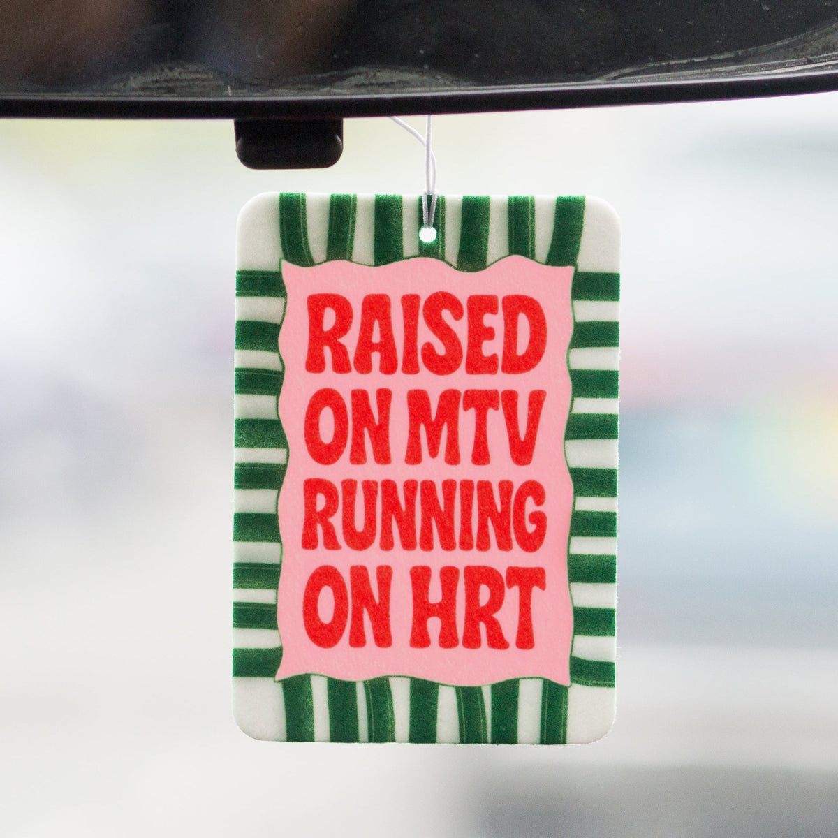 Car mirror with a decorative air freshener reading 'Raised on MTV Running on HRT' with green and white stripes.