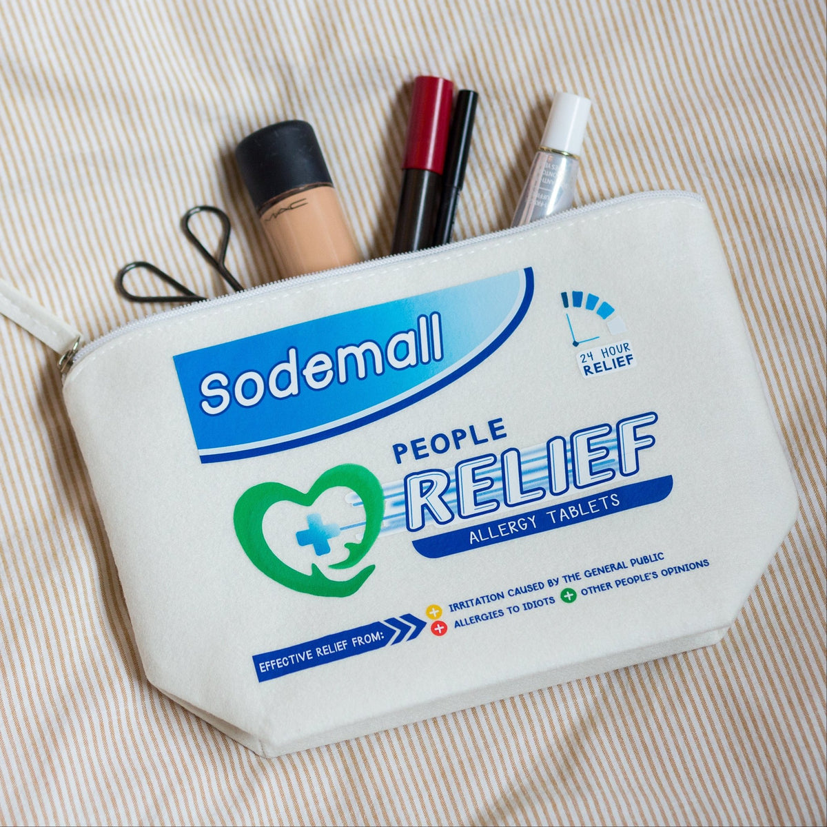 A cream-colored canvas zippered pouch printed with 'Sodemall People Relief Allergy Tablets' branding, filled with various personal care items, laid flat on a beige and white striped fabric background.