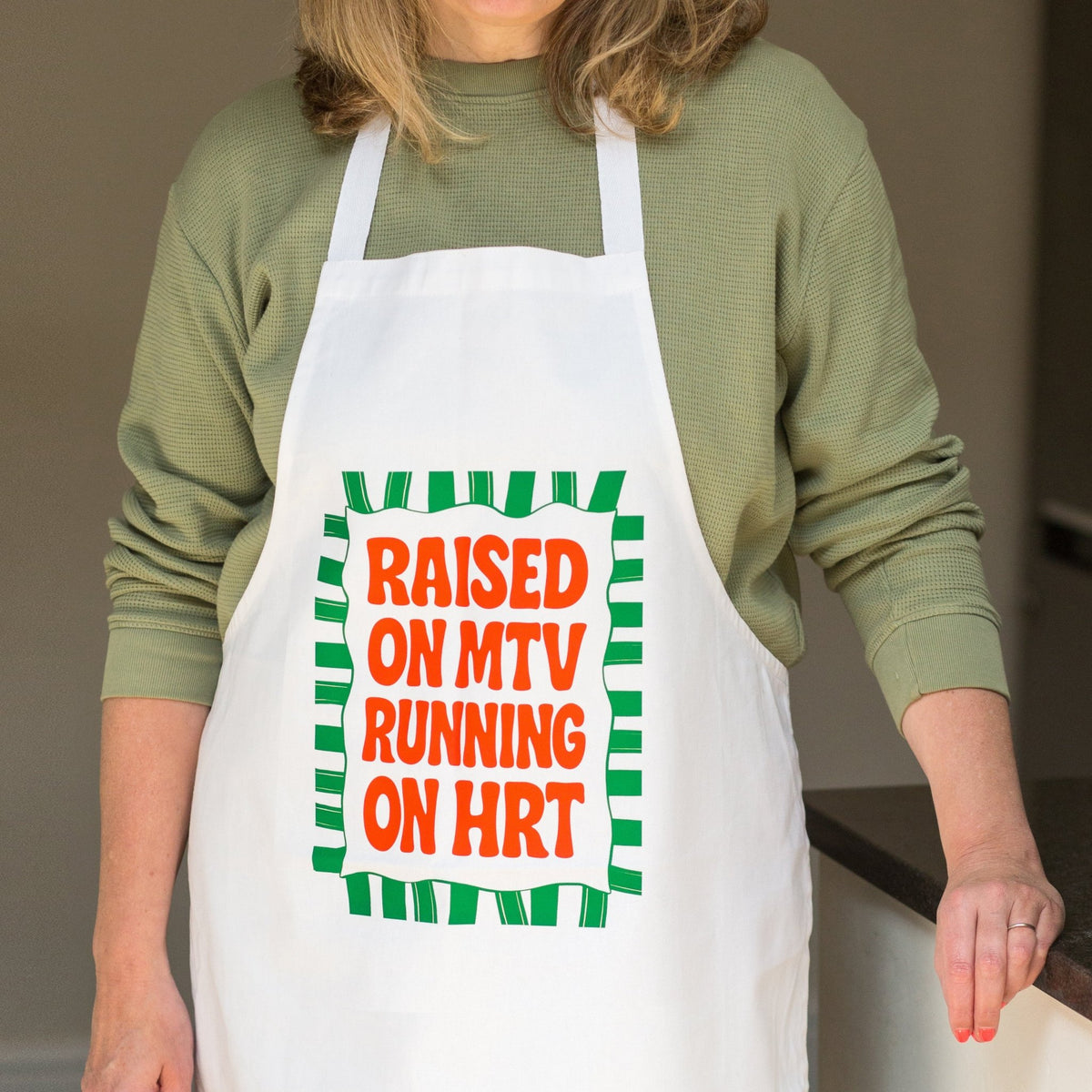 A person wearing a white apron with a green and orange graphic that reads 'RAISED ON MTV RUNNING ON HRT' over a sage green long-sleeve shirt, photographed indoors.