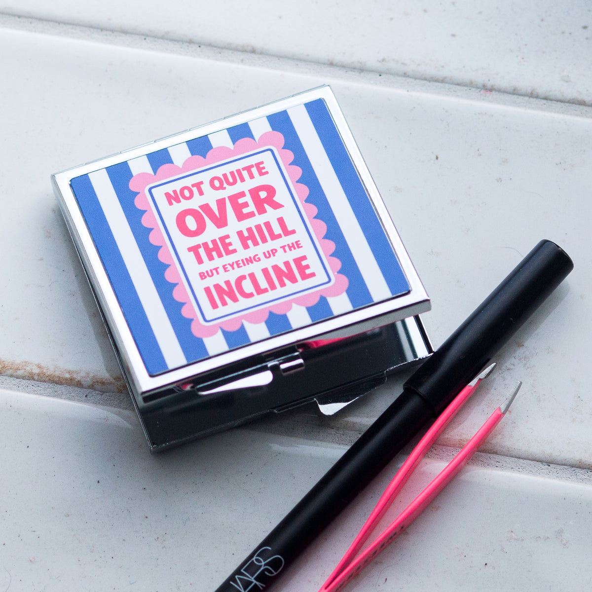 Compact mirror with humorous text and a pen on a light wooden surface