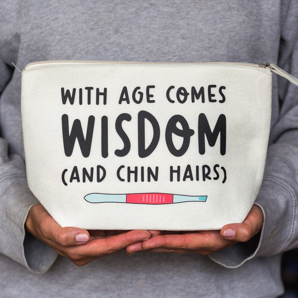 A person in a gray sweatshirt holds a cream-colored zippered pouch with the text 'WITH AGE COMES WISDOM (AND CHIN HAIRS)' and a small pink and blue pregnancy test graphic.