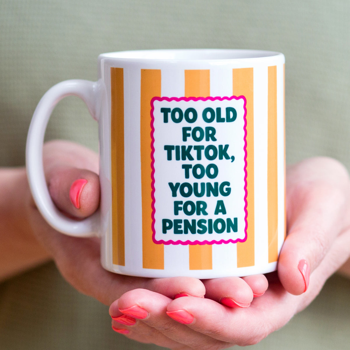 Mug with text 'Too old for Tiktok, too young for a pension' held by hands with red nail polish.
