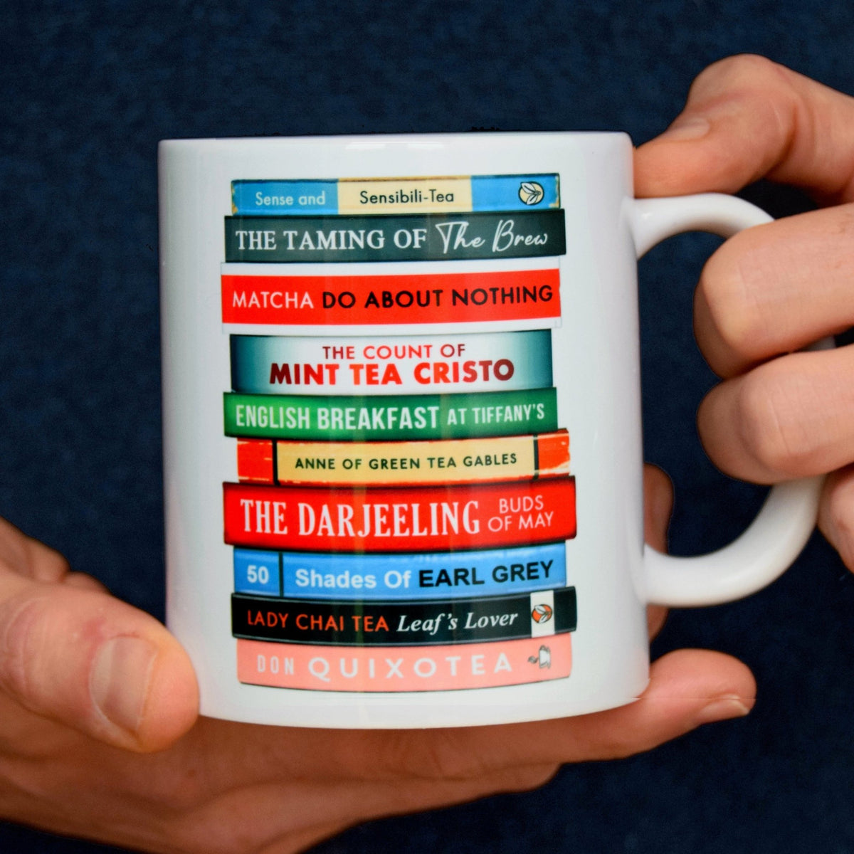 Mug with book stack featuring titles that are tea themed puns on famous novels, held by hands against a dark background
