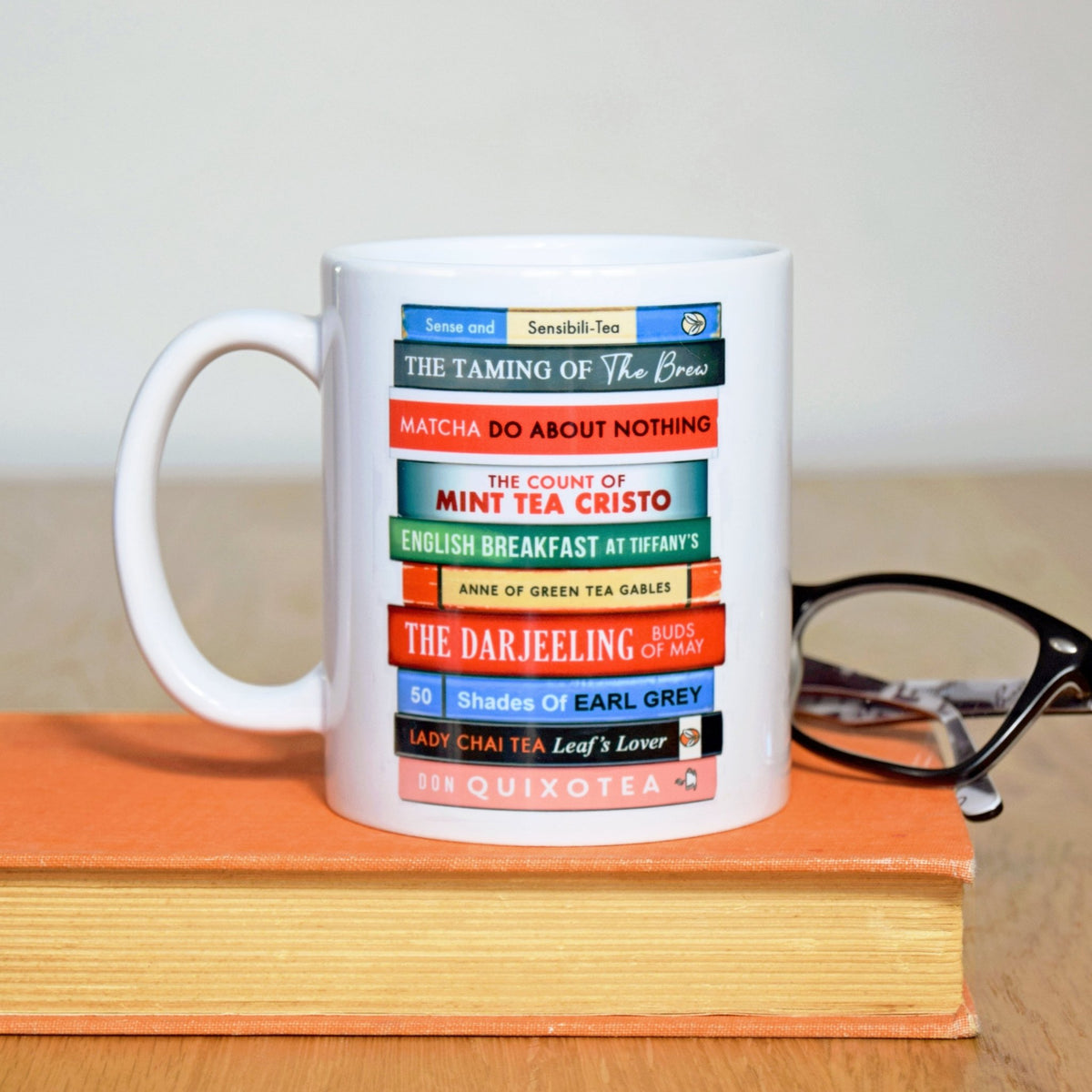 White mug with colorful text design on a wooden surface with a book and glasses.