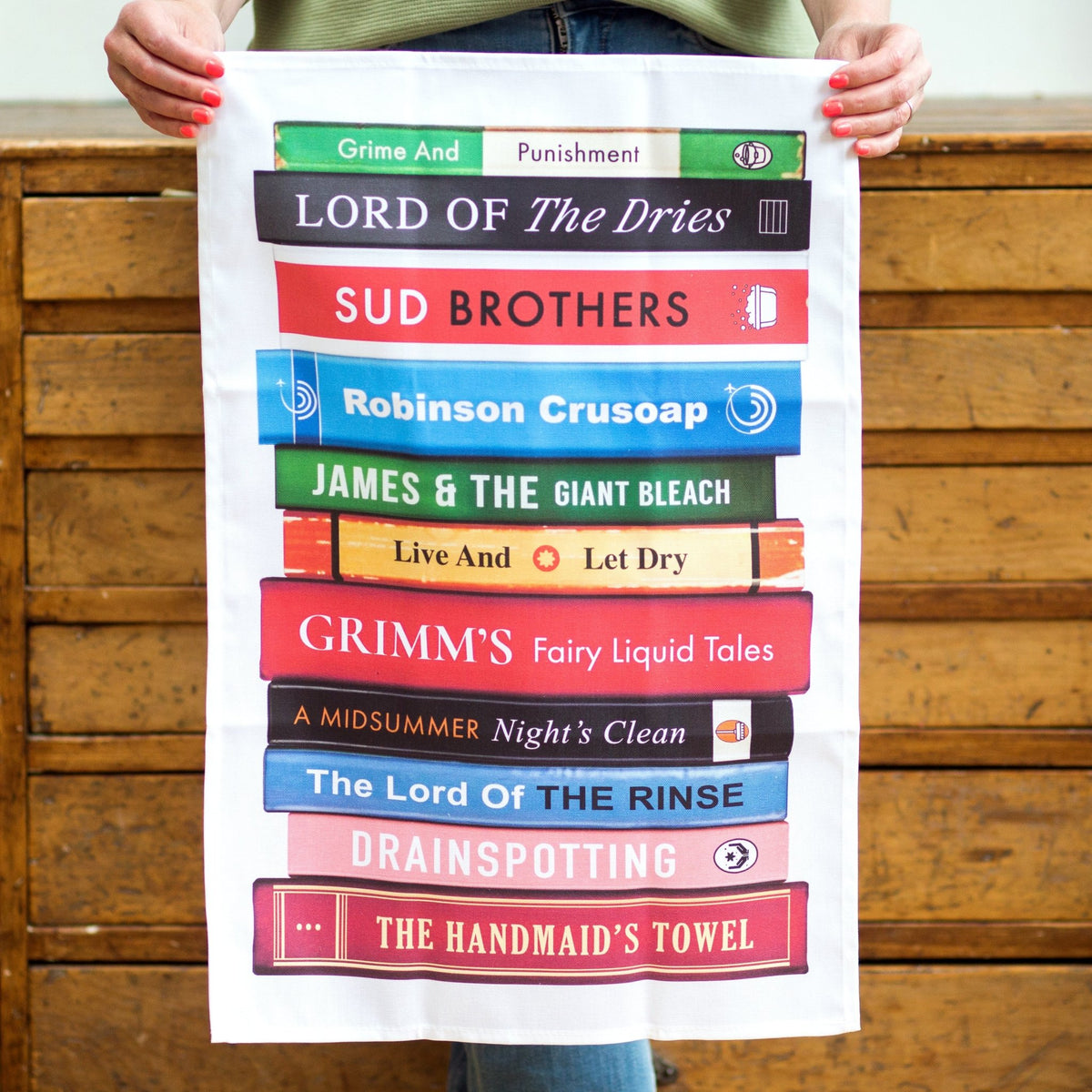 tea towel with washing up themed puns on book titles held in hands against a wooden background