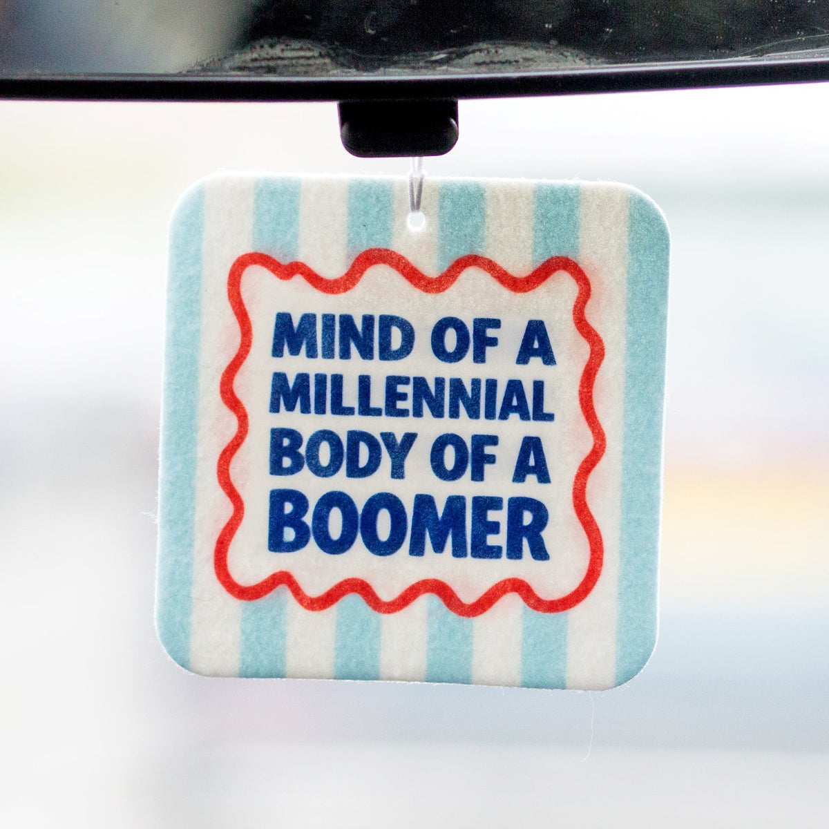 Car air freshener with text 'Mind of a Millennial, Body of a Boomer' on a blue and white striped background.