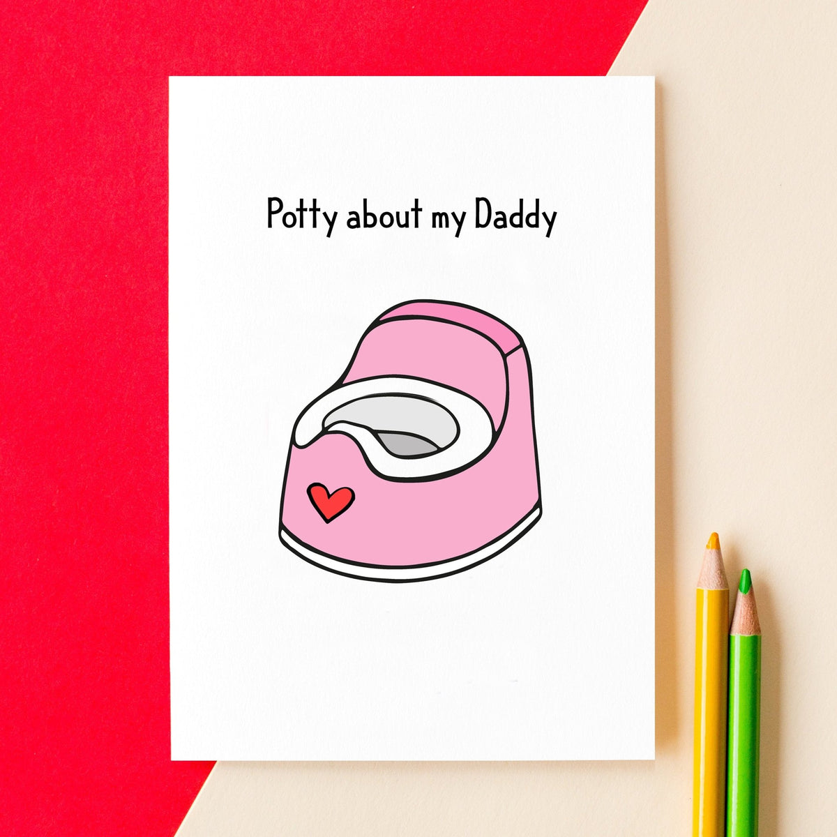 A white greeting card with the text 'Potty about my Daddy' and a pink potty illustration, styled on a beige surface with a red paper backdrop and two colored pencils.