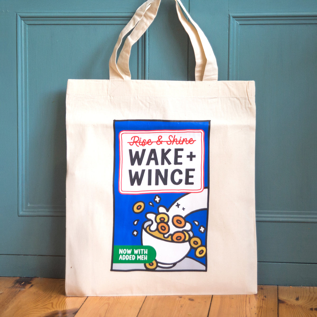 A cream canvas tote bag with a printed graphic of a cereal box labeled 'Ripe & Shire WAKE + WINCE' hanging against a teal paneled door on a wooden floor.