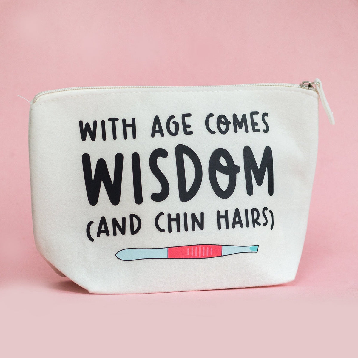 An off-white canvas zippered pouch with black text reading 'WITH AGE COMES WISDOM (AND CHIN HAIRS)' and a small pink and blue pregnancy test graphic, set against a solid pink background.