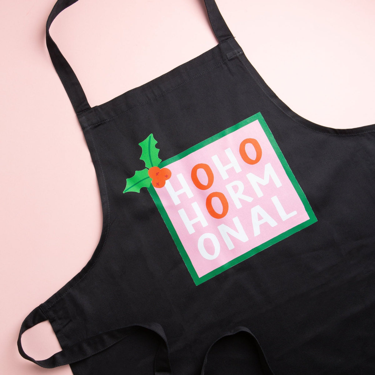 a close up of a black apron that says hohohormonal lying flat on a pink background