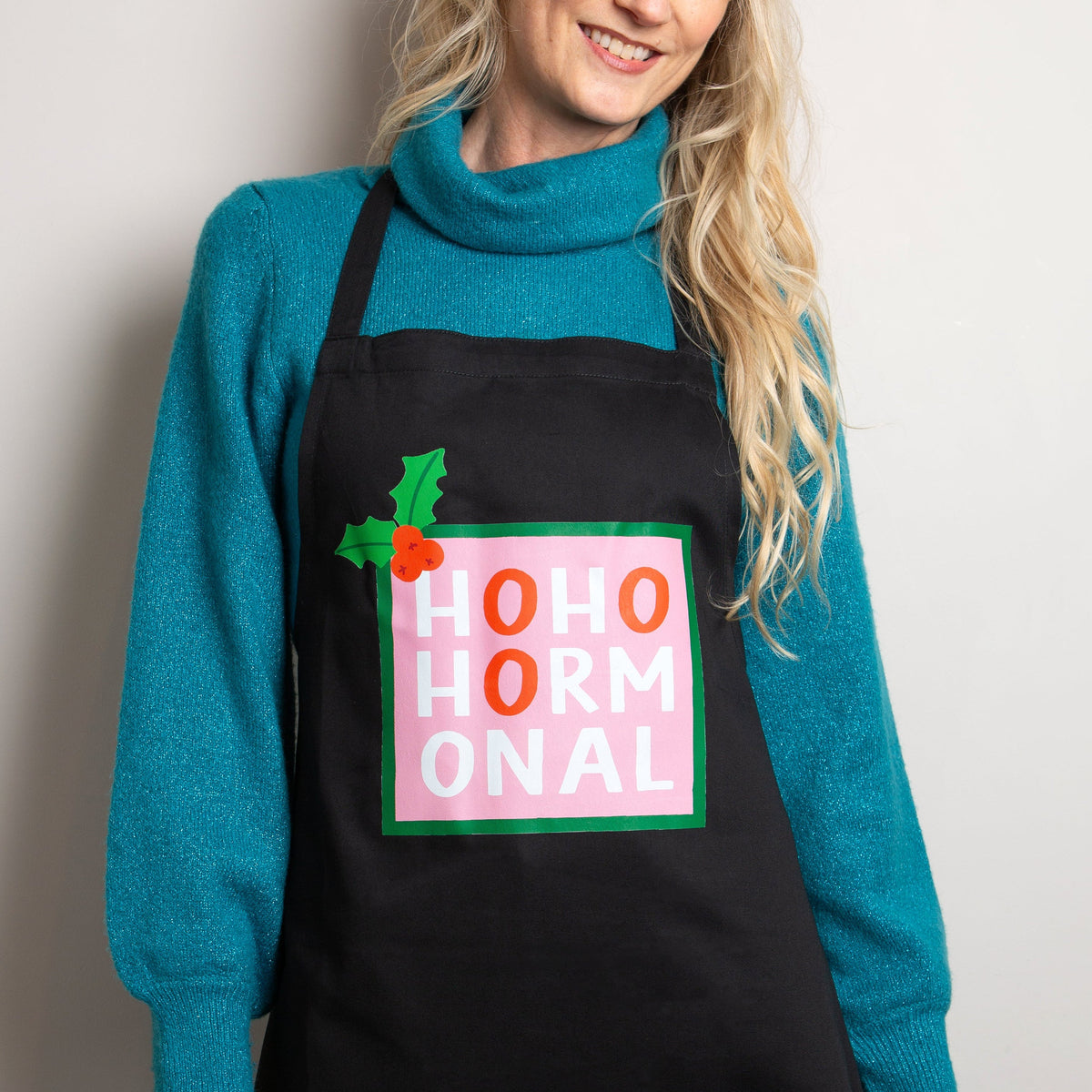 female wearing a blue jumper and black apron that says 'HoHoHormonal' with an illustrated sprig of holly