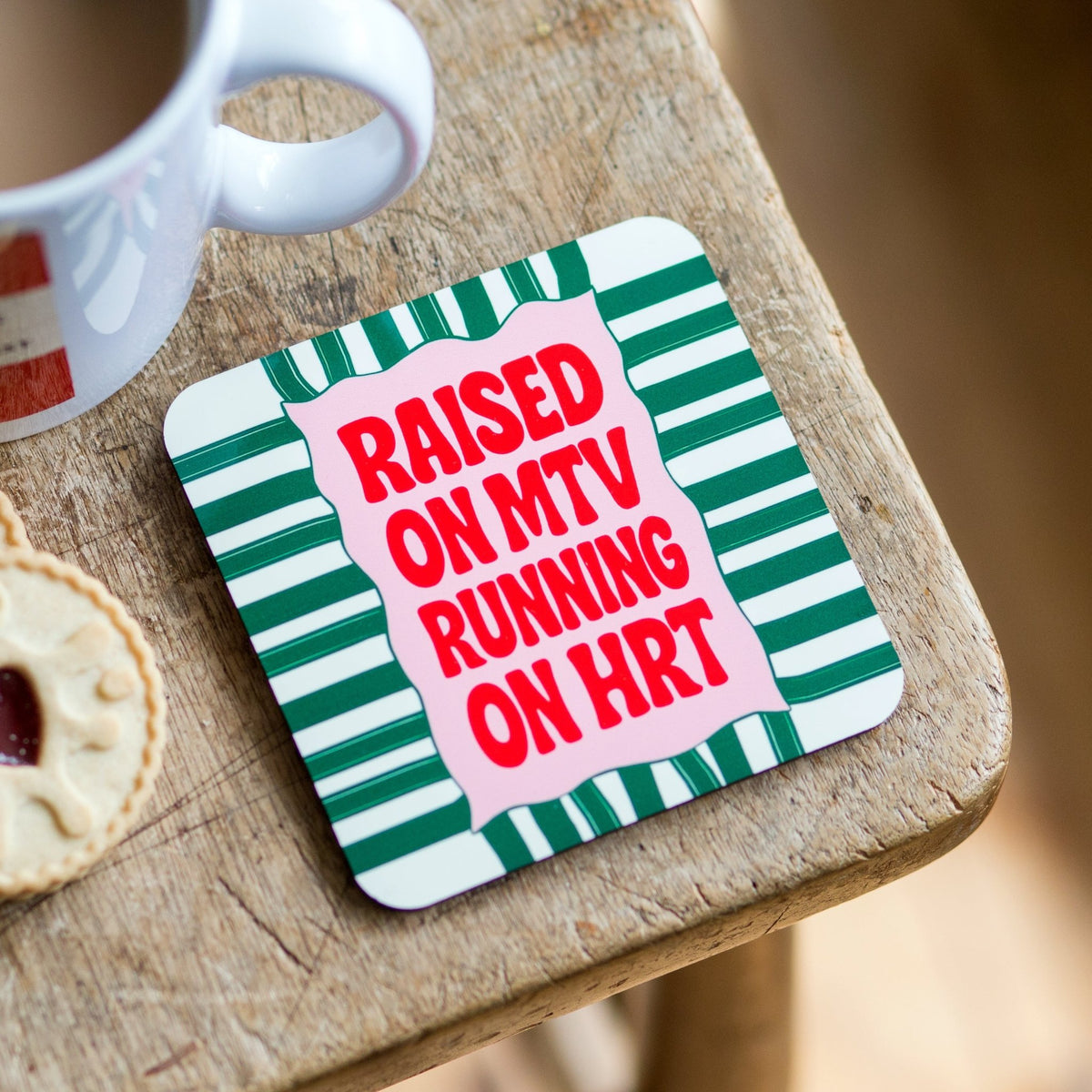 Coaster with 'Raised on MTV Running on HRT' text on a wooden surface with a cup and cookie.