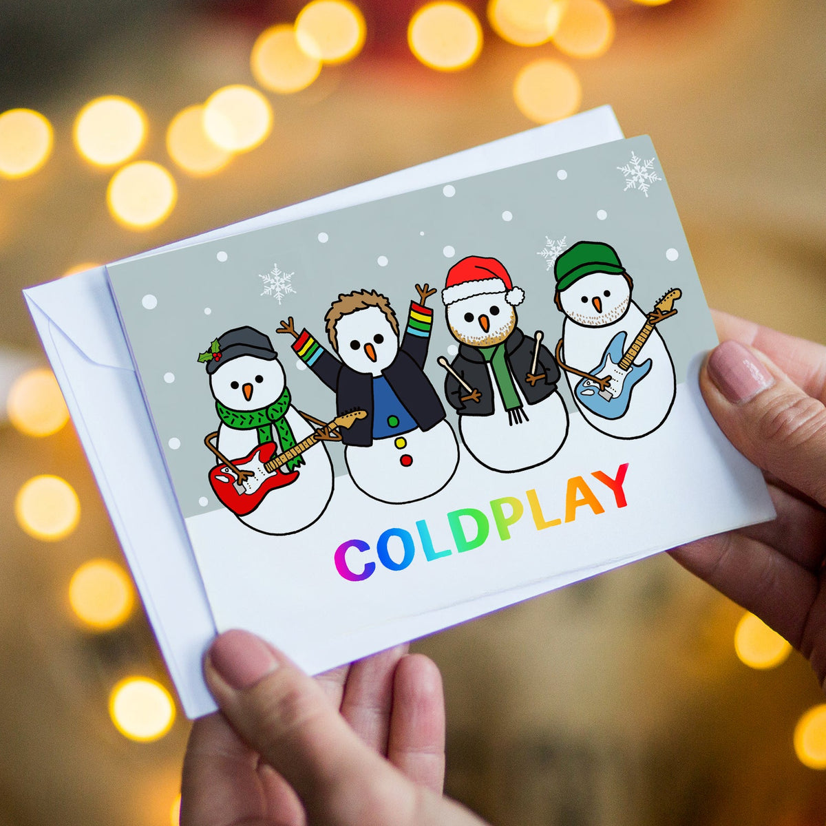 A person holds a Christmas Christmas card featuring four cartoon snowmen playing music, with the word 'COLDPLAY' in rainbow letters. The card is shown with a white envelope against a warm, blurred background of bokeh holiday lights.