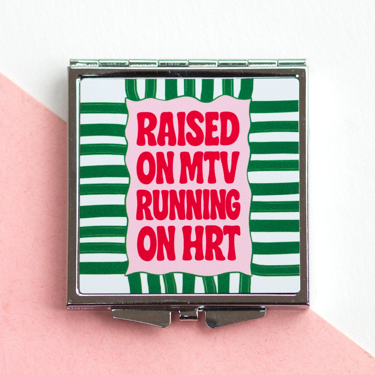 Compact mirror with green and white striped design and red text on a pink surface.