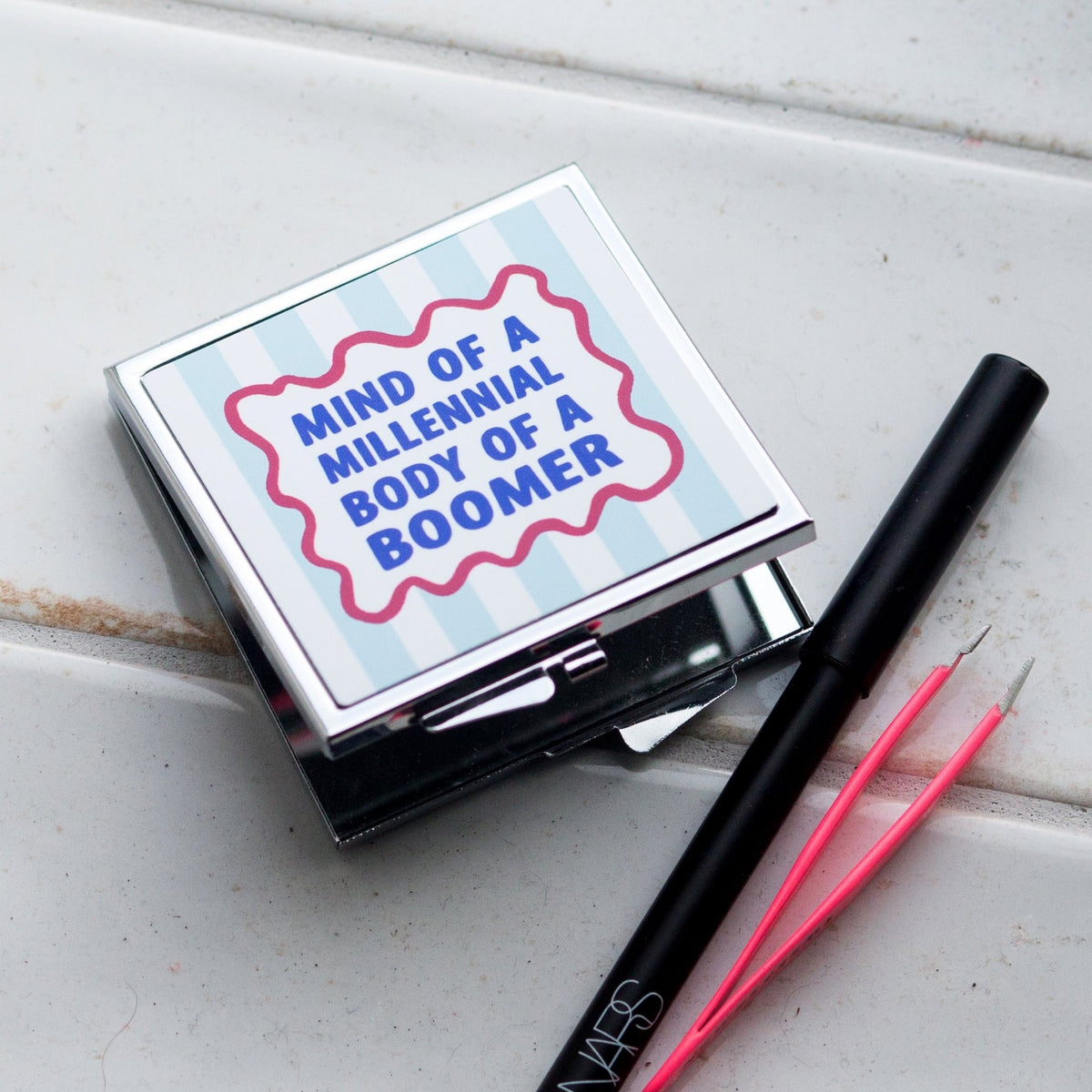 Compact mirror with humorous text on a white surface with makeup tools.