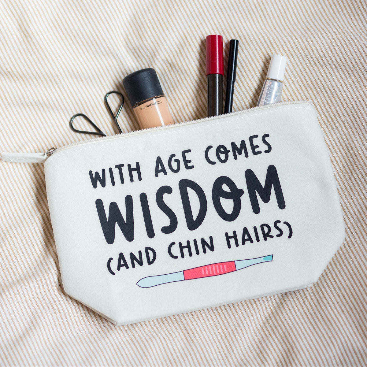 An off-white canvas cosmetic pouch with black text reading 'WITH AGE COMES WISDOM (AND CHIN HAIRS)' and a small pregnancy test graphic, shown on a beige striped fabric background with makeup items peeking out from the top.