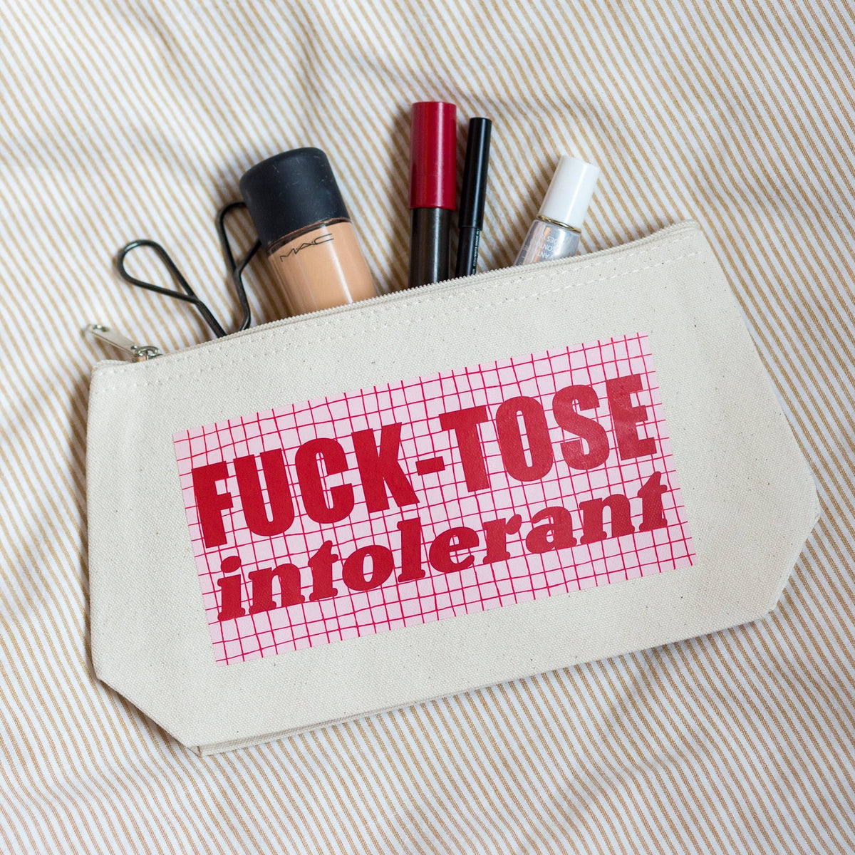A beige canvas makeup pouch with a pink grid pattern and the text 'FUCK-TOSE intolerant' printed on the front, shown with makeup items inside on a striped fabric background.