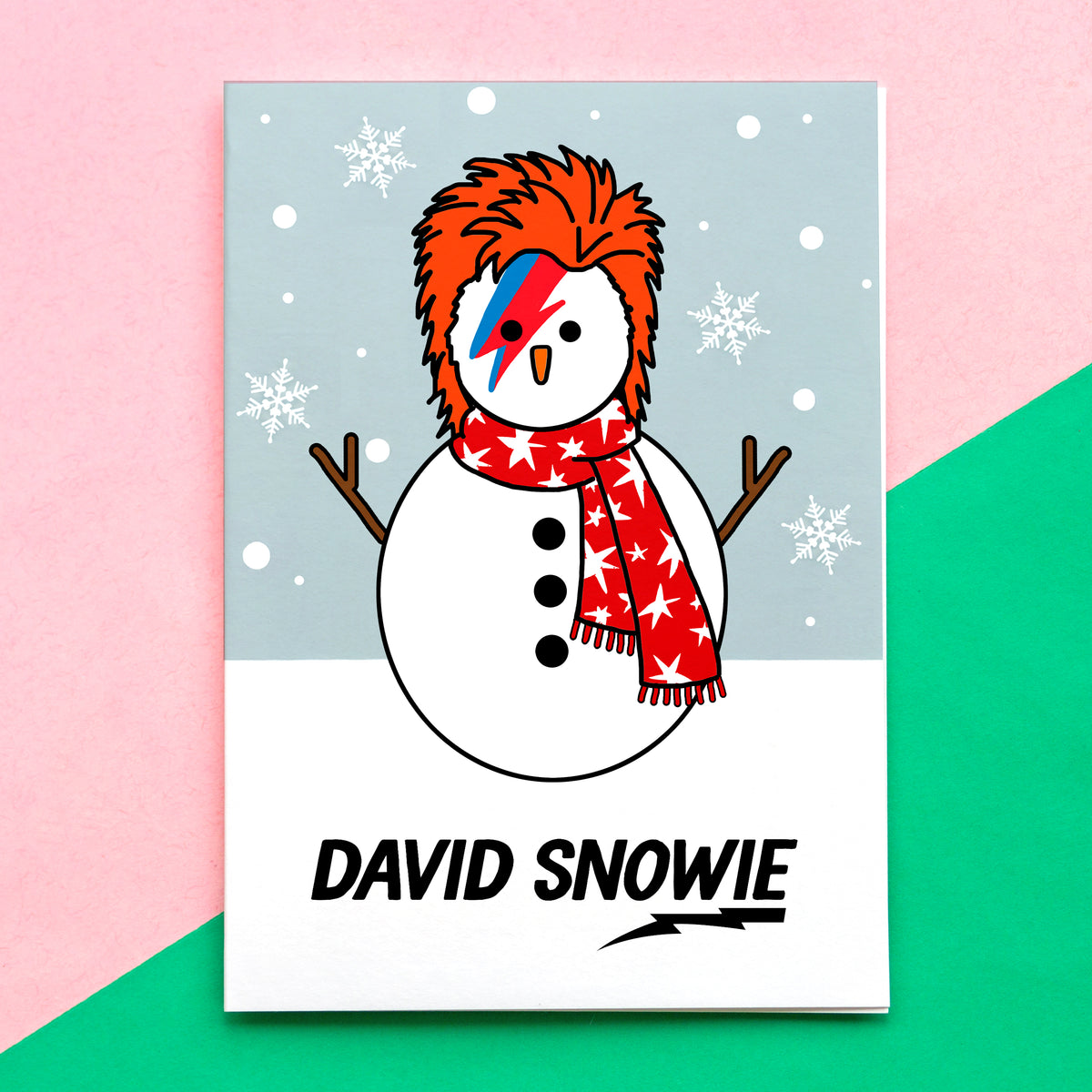 A greeting card featuring a cartoon snowman styled like David Bowie, with orange hair, a red-and-blue lightning-embellished face, and a red star-patterned scarf. The card is shown standing upright against a split pink and green background.