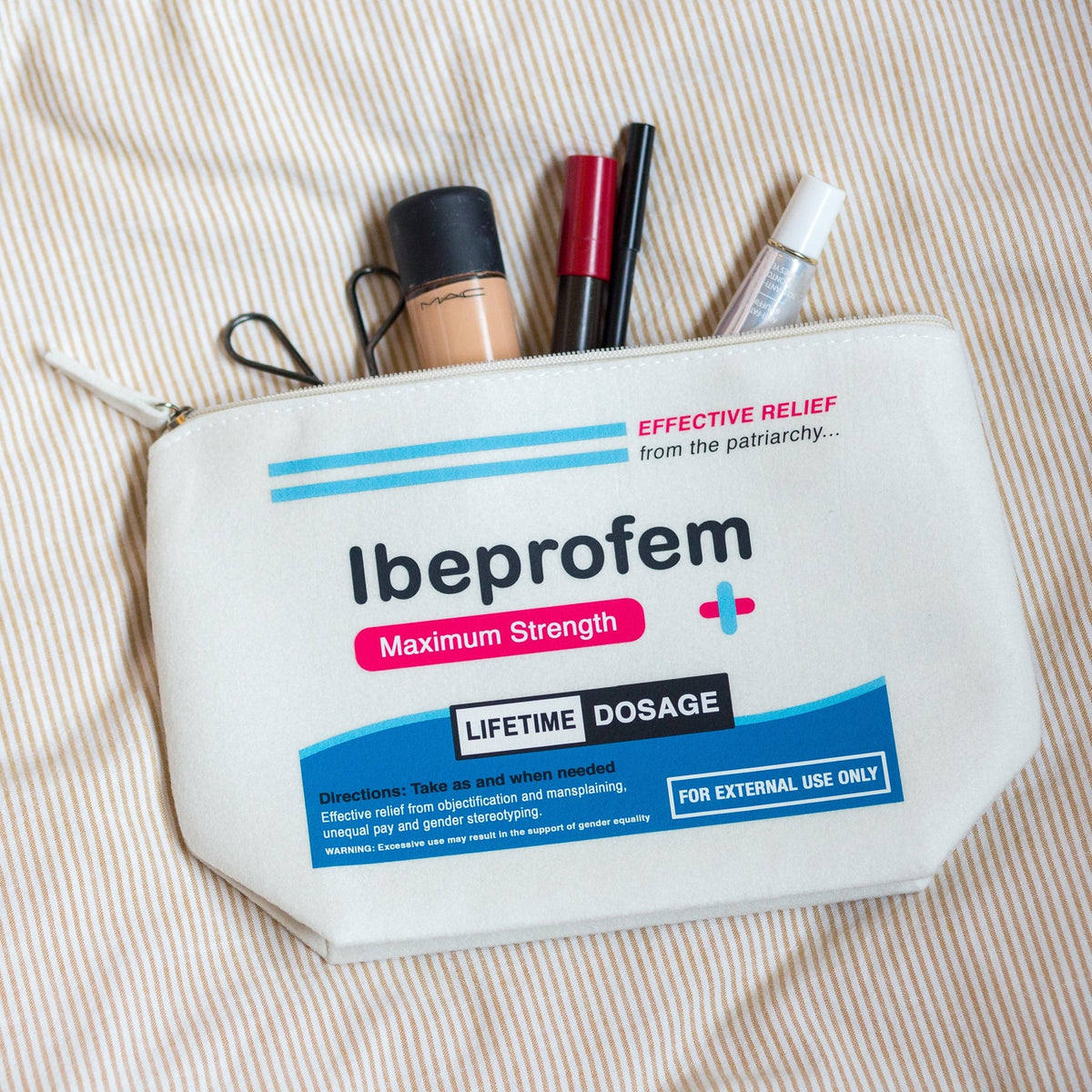 A white zippered pouch labeled 'Ibuprofen Maximum Strength' with makeup items peeking out from the top, placed on a beige-and-white striped fabric background.