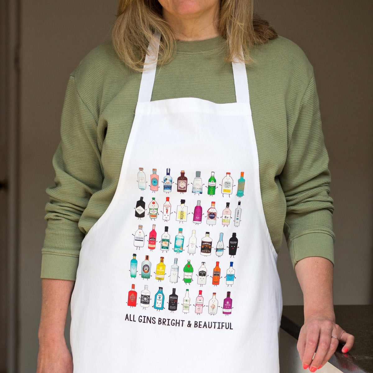 A woman wearing a white apron with a colorful grid of cocktail and spirit bottle illustrations and the text 'ALL GINS BRIGHT & BEAUTIFUL' over an olive green long-sleeve shirt.