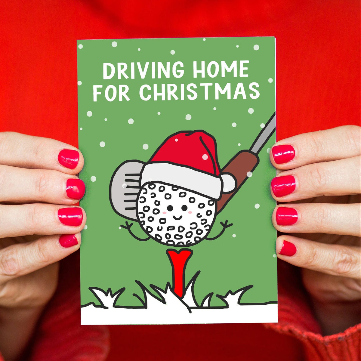 A person holding a green Christmas card with the text 'DRIVING HOME FOR CHRISTMAS' and a cartoon golf ball wearing a Santa hat and red sweater background.