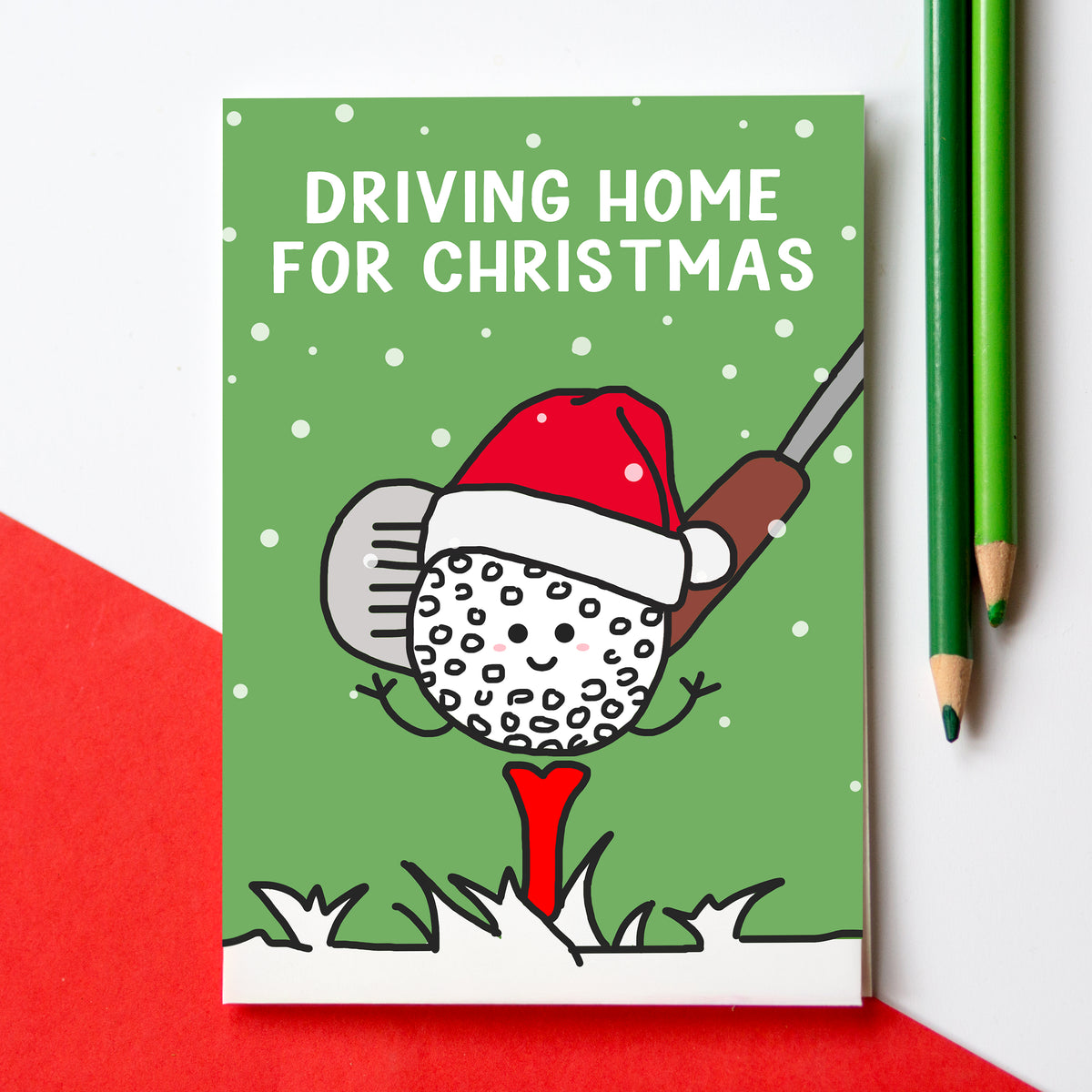 A Christmas card with a green background and the text 'DRIVING HOME FOR CHRISTMAS' at the top. The card features a cartoon golf ball wearing a red Santa hat and using a golf club to lift a red Christmas tree.