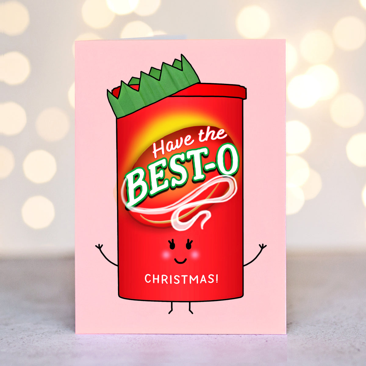 A pink greeting card featuring a cartoon red candy cane with green trim and the text 'Have the BEST-O CHRISTMAS!' on the front, photographed against a softly blurred warm bokeh background.
