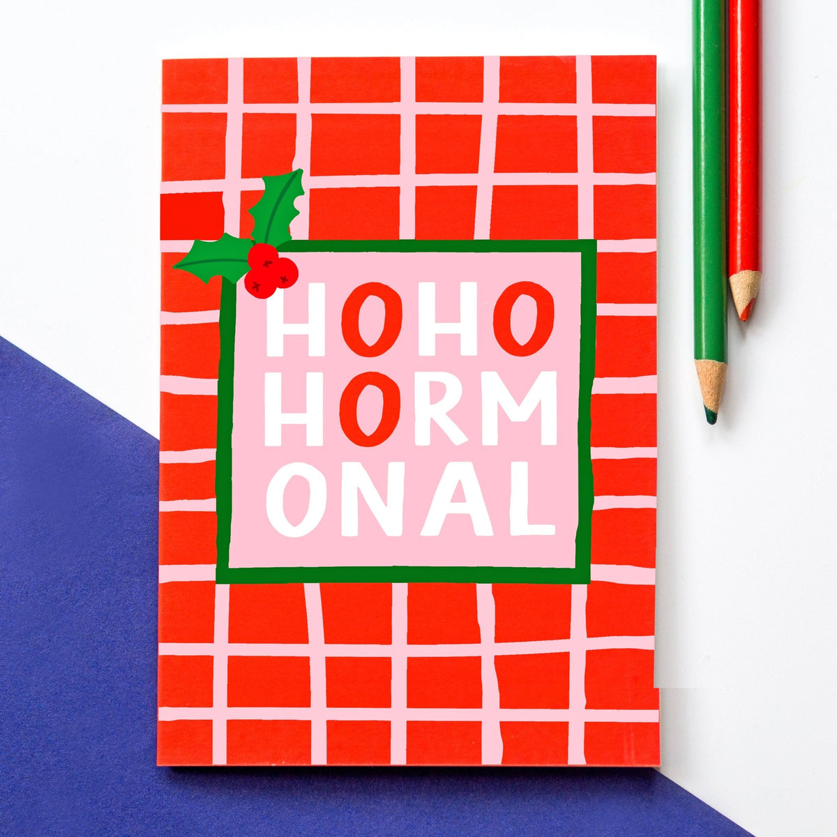 A Christmas card with a festive red and white grid pattern, featuring a central pink panel with the text 'HOHO HORMONAL' in bold red and white letters, decorated with a small holly sprig. The card is photographed on a white surface with a blue corner and two colored pencils nearby.