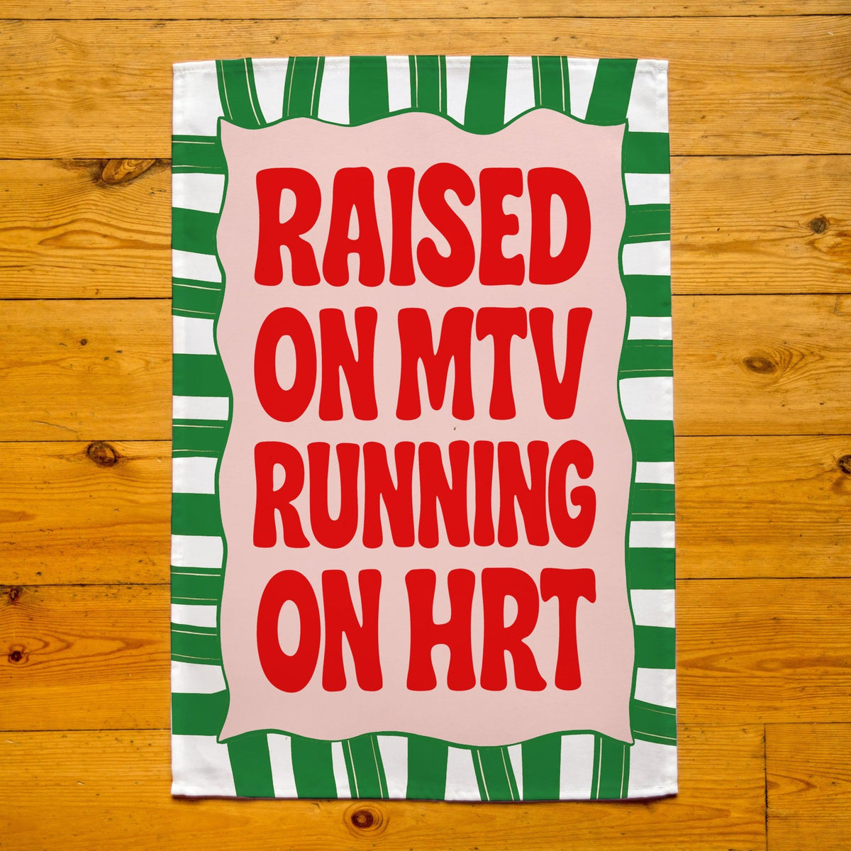 Tea towel with text 'Raised on MTV Running on HRT' on a wooden surface
