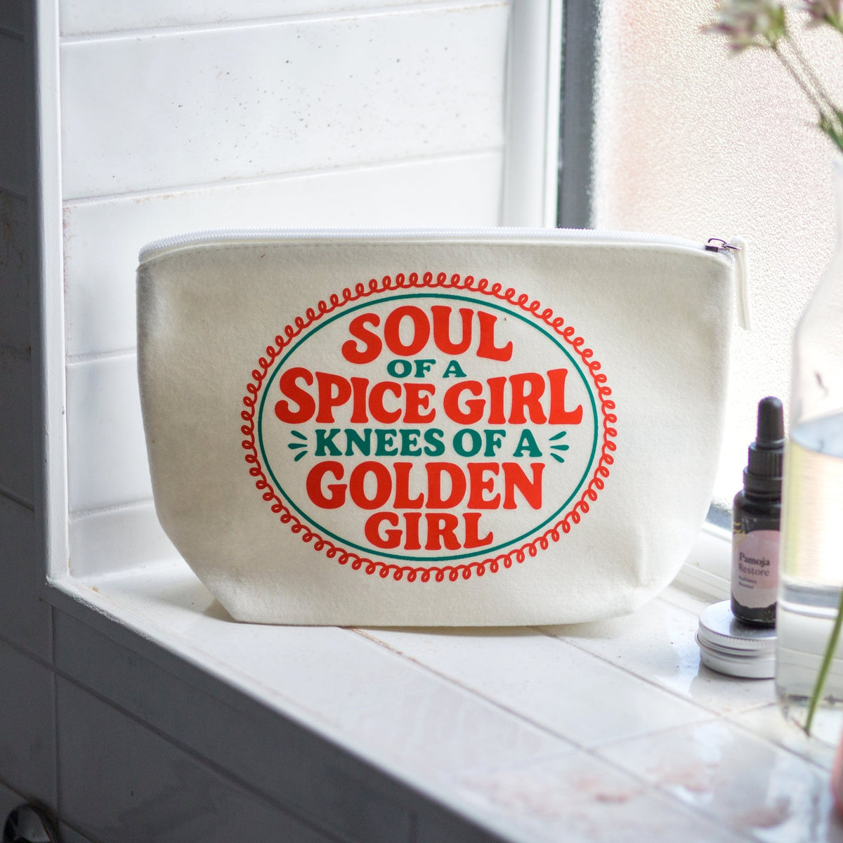 An off-white canvas cosmetic pouch with a colorful printed graphic reading 'SOUL OF A SPICE GIRL KNEES OF A GOLDEN GIRL' sits on a white tiled windowsill next to a small amber dropper bottle.