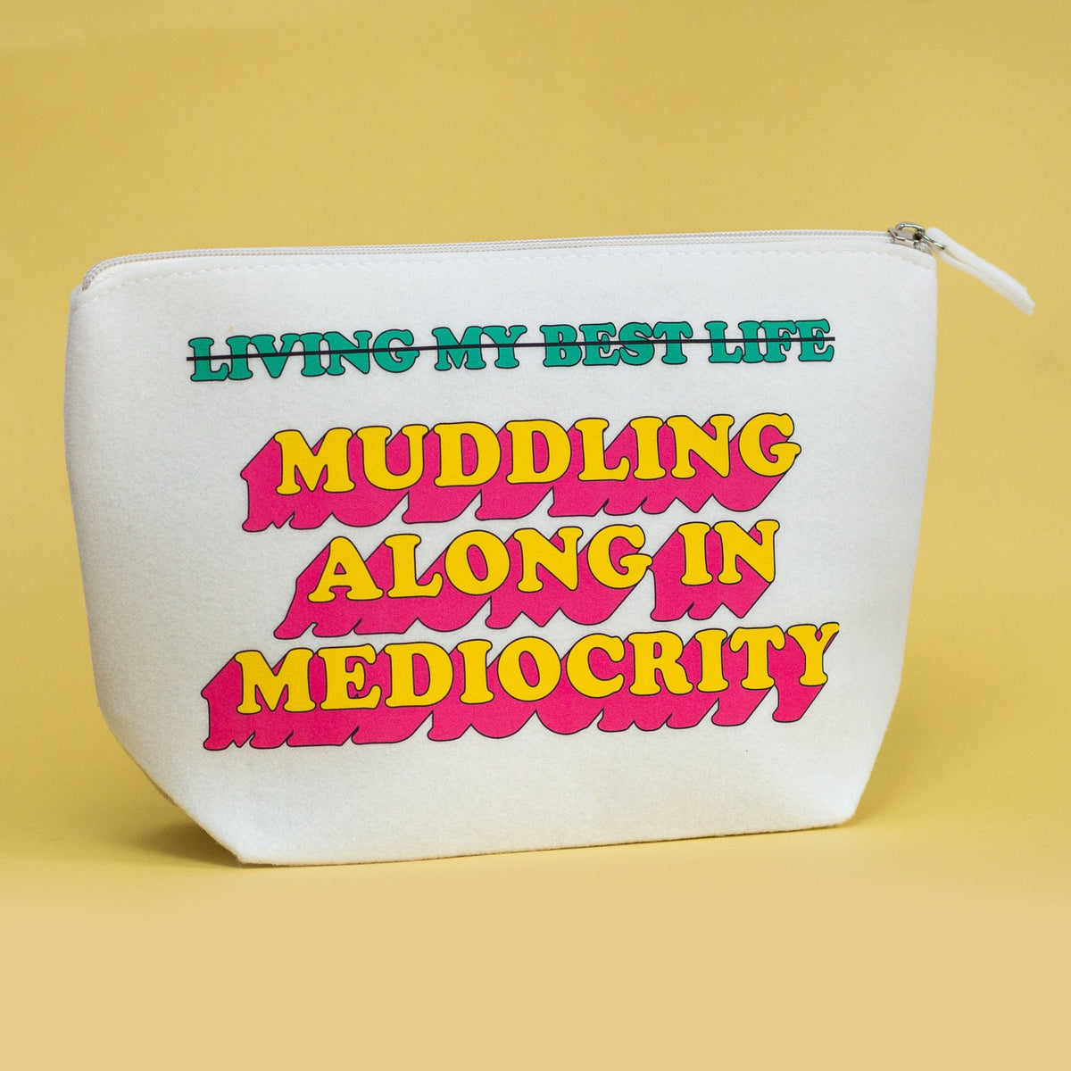 A white zippered pouch with bold retro-style text reading 'LIVING MY BEST LIFE Muddling ALONG IN MEDIACRITY' in green and yellow with pink shadowing, set against a solid pale yellow background.