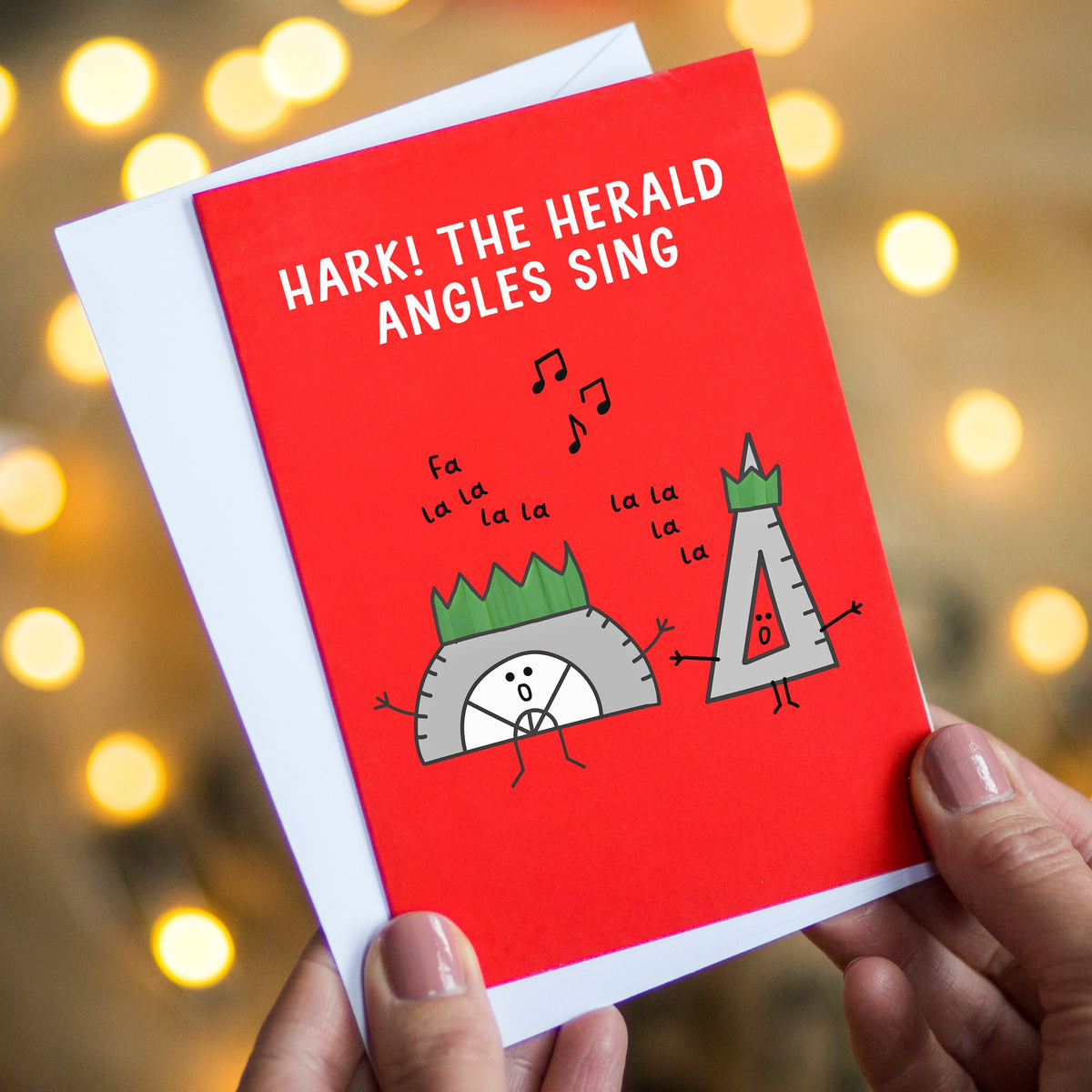 A person holds a red greeting card with a white envelope behind it. The card has the text 'HARK! THE HERALD ANGLES SING' and cartoon illustrations of two angles singing.