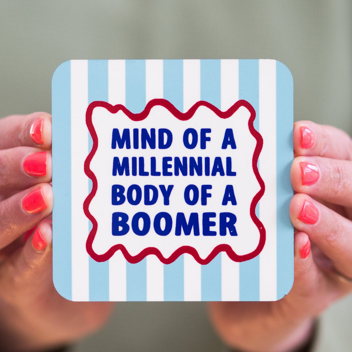 Coaster with 'Mind of a Millennial, Body of a Boomer' text held by hands with red nail polish.