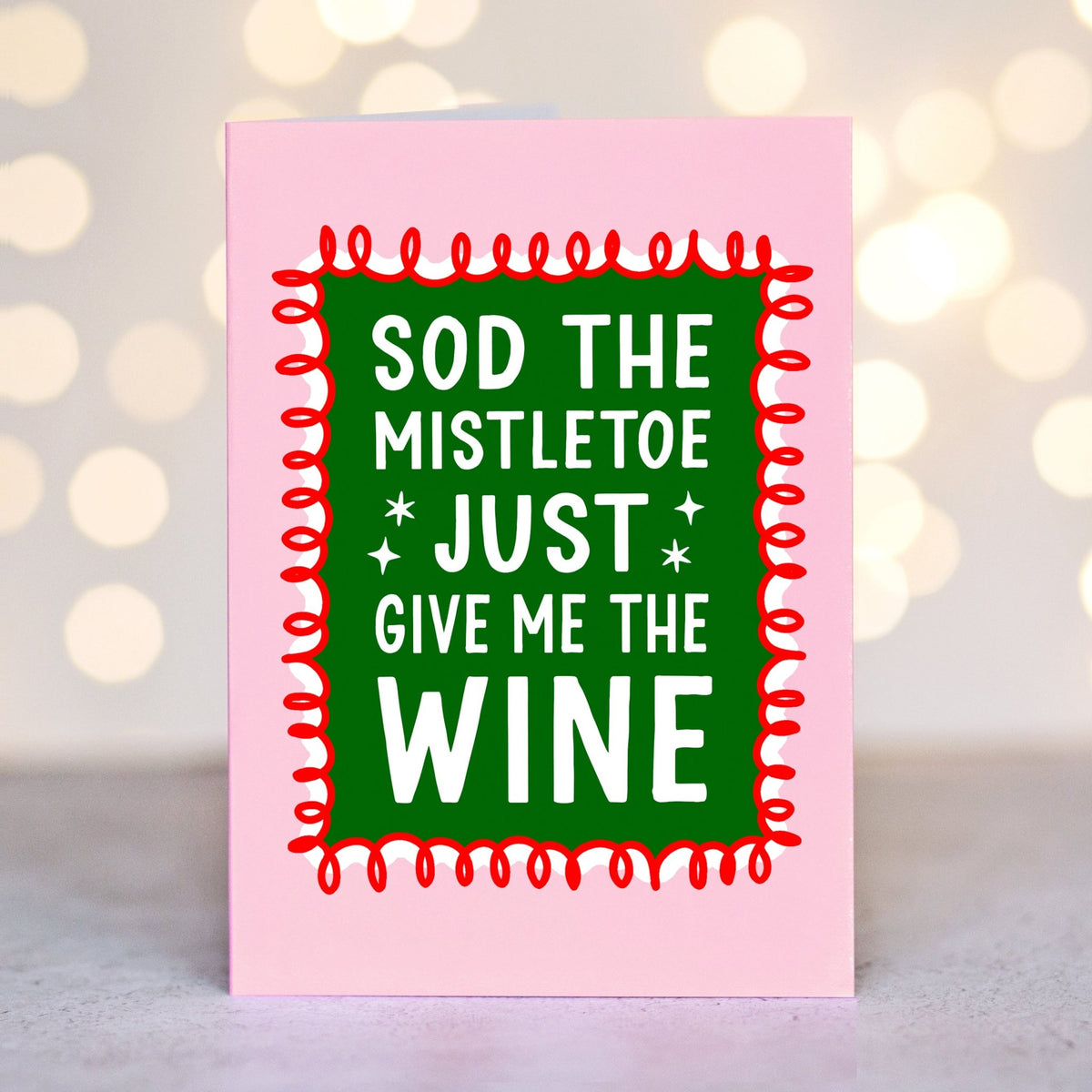 A pink christmas card with a green central panel and bold white text reading 'SOD THE MISTLETOE JUST GIVE ME THE WINE', framed by a red zigzag border. The card is photographed upright against a softly blurred warm bokeh background.